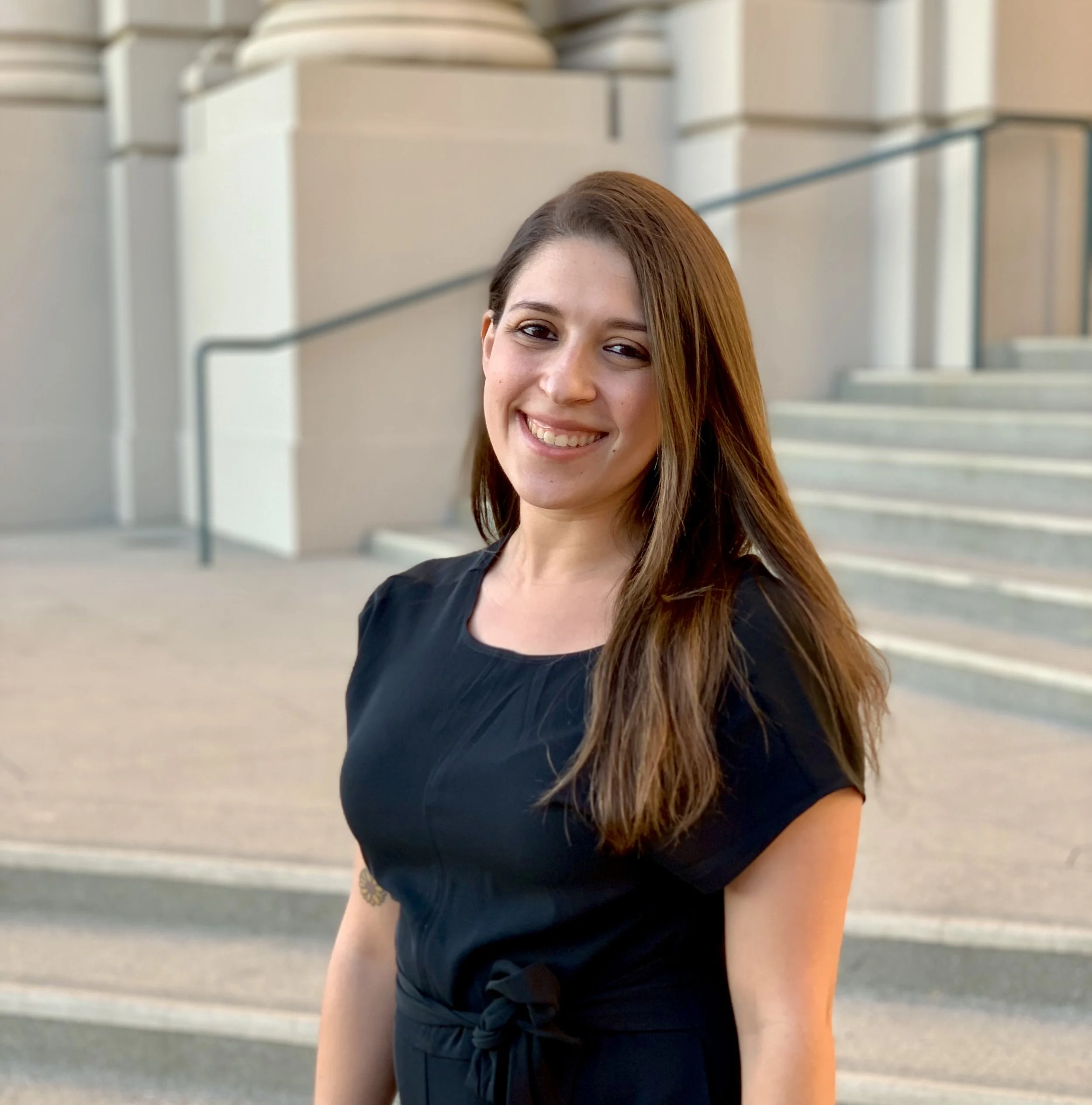 A young woman with long brown hair, smiling, wearing a black dress, standing outdoors near stairs and a building.