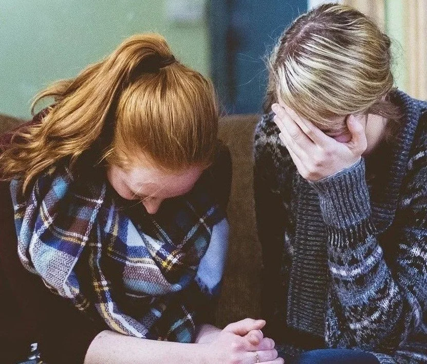 Two women sitting on a couch, holding hands, with their heads bowed and hands covering their faces, appearing upset or distressed.