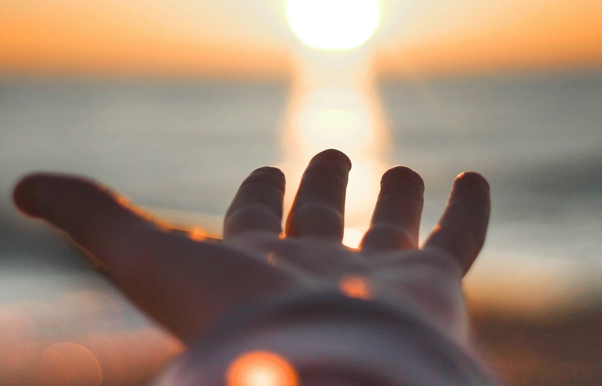 A hand reaches out towards the setting sun over a body of water.