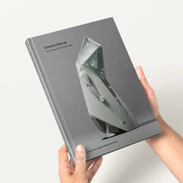Person holding a gray owner’s manual titled 'The Catalog of 2022 Technologies' with a futuristic vehicle illustration on the cover.