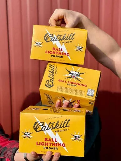 Person holding a stack of three yellow boxes of Catskill Brewery Ball Lightning Pilsner beer, with a pink wall in the background.