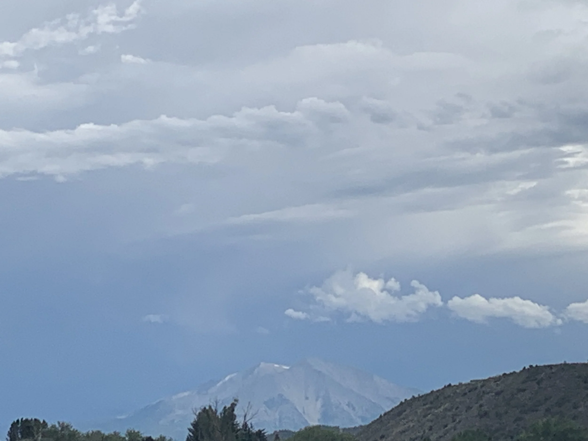 A mountainous landscape with a cloudy sky overhead. The mountain peaks are visible in the distance, with a layer of clouds partially covering them. The foreground shows a hill with trees.