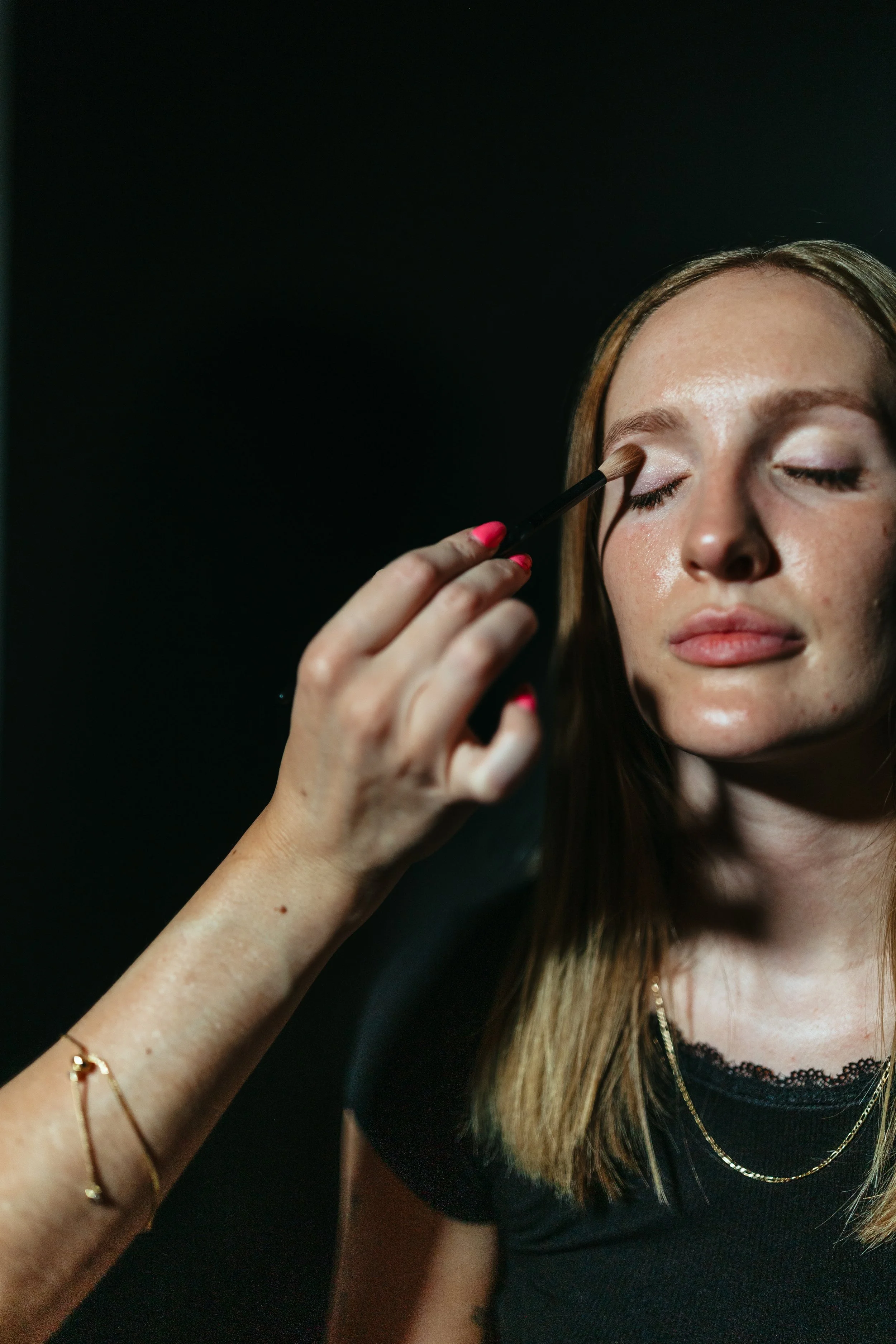 A woman with light brown hair and fair skin applying eyeshadow with a brush while wearing a black top and layered gold necklaces, against a dark background.