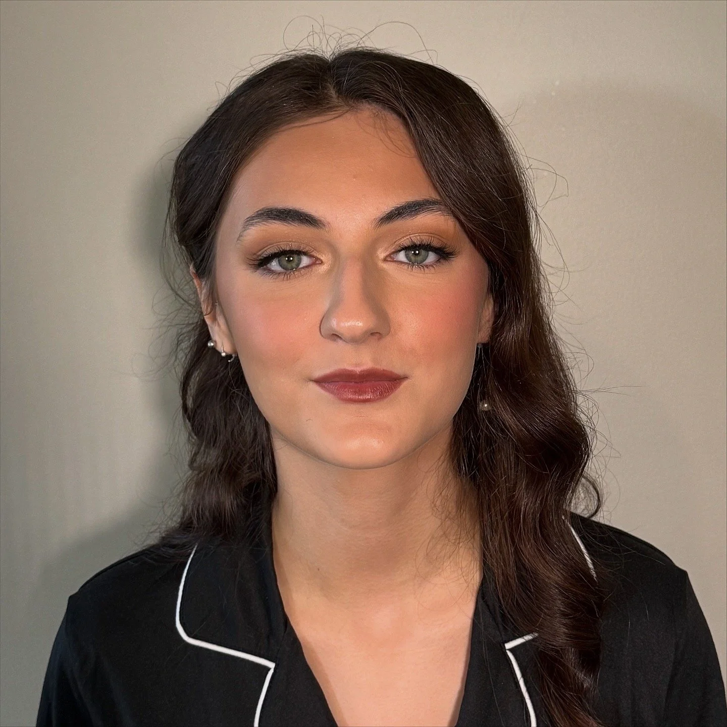 Close-up portrait of a young woman with brown hair, green eyes, and makeup, wearing a black shirt with white piping, against a neutral background.