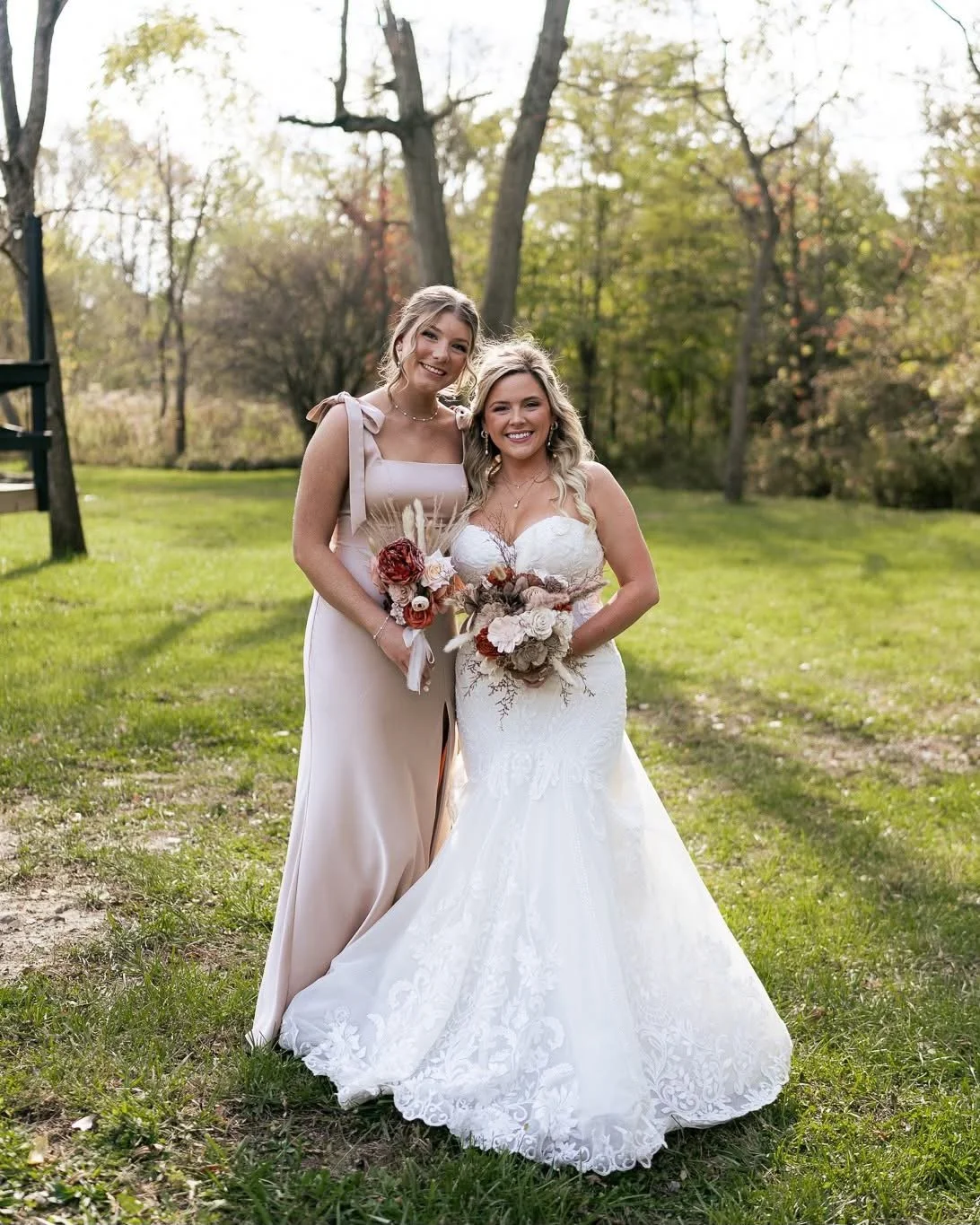Two women, one in a white wedding dress and the other in a light pink dress, standing outdoors on grass with trees in the background, smiling and holding bouquets of flowers.