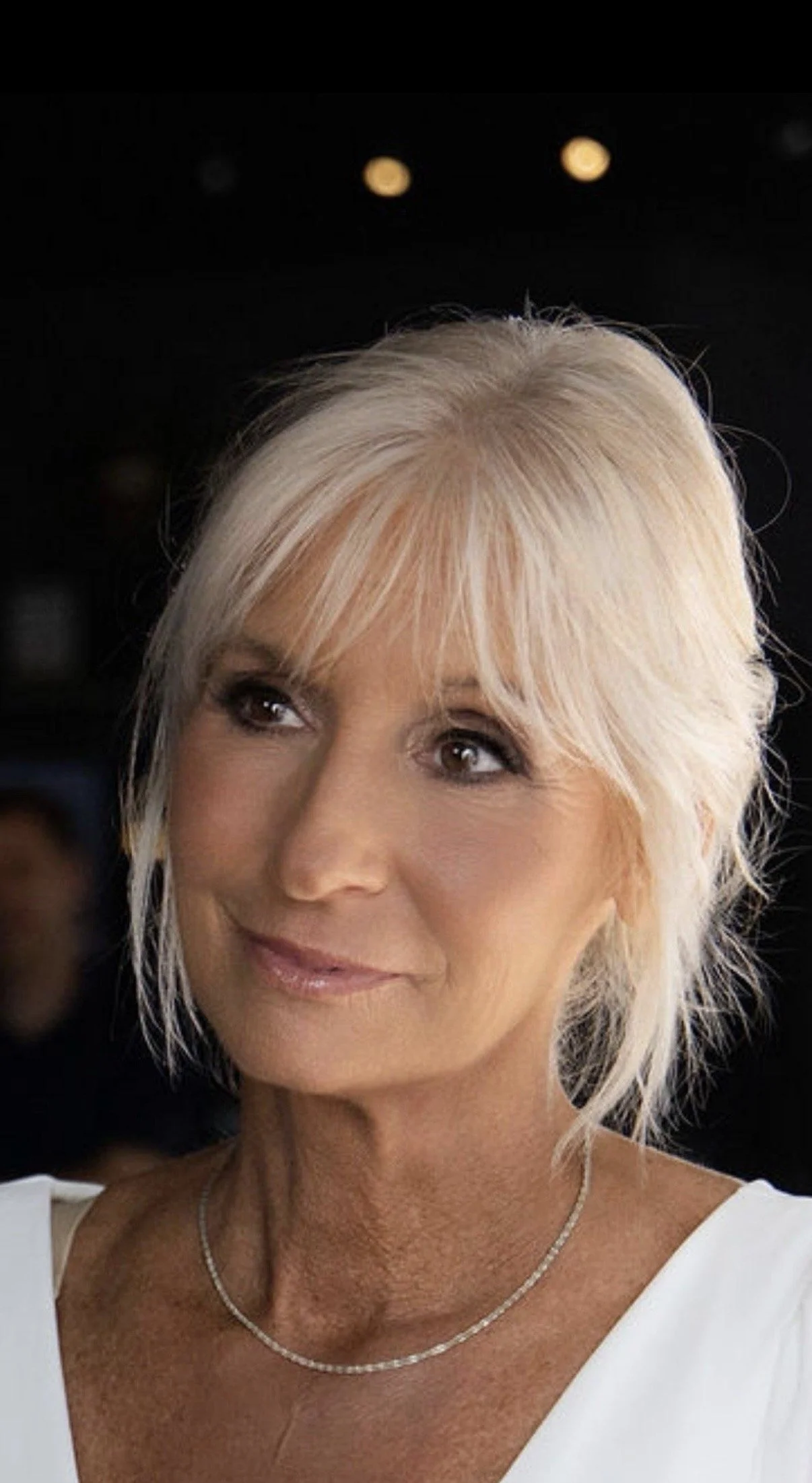 A close-up portrait of an older woman with blonde hair styled in a layered cut, wearing a white top and a delicate silver necklace, smiling slightly with a dark background.
