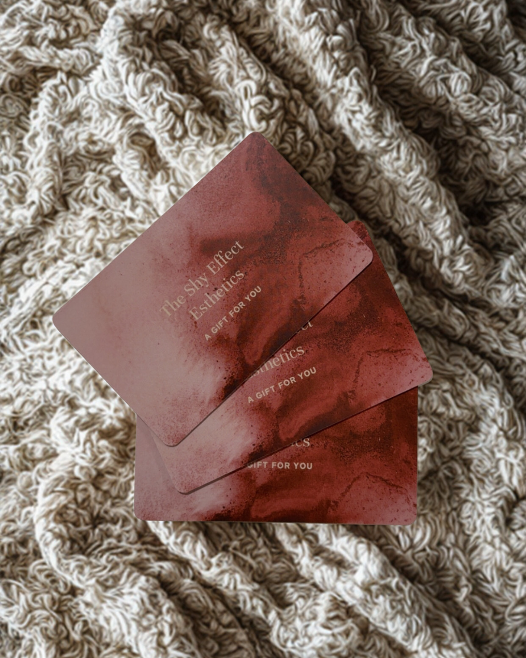 Three pink cards with abstract red and black designs on a textured, beige knitted blanket. The cards are labeled "The SHY Effect Esthetics" and "A GIFT FOR YOU."