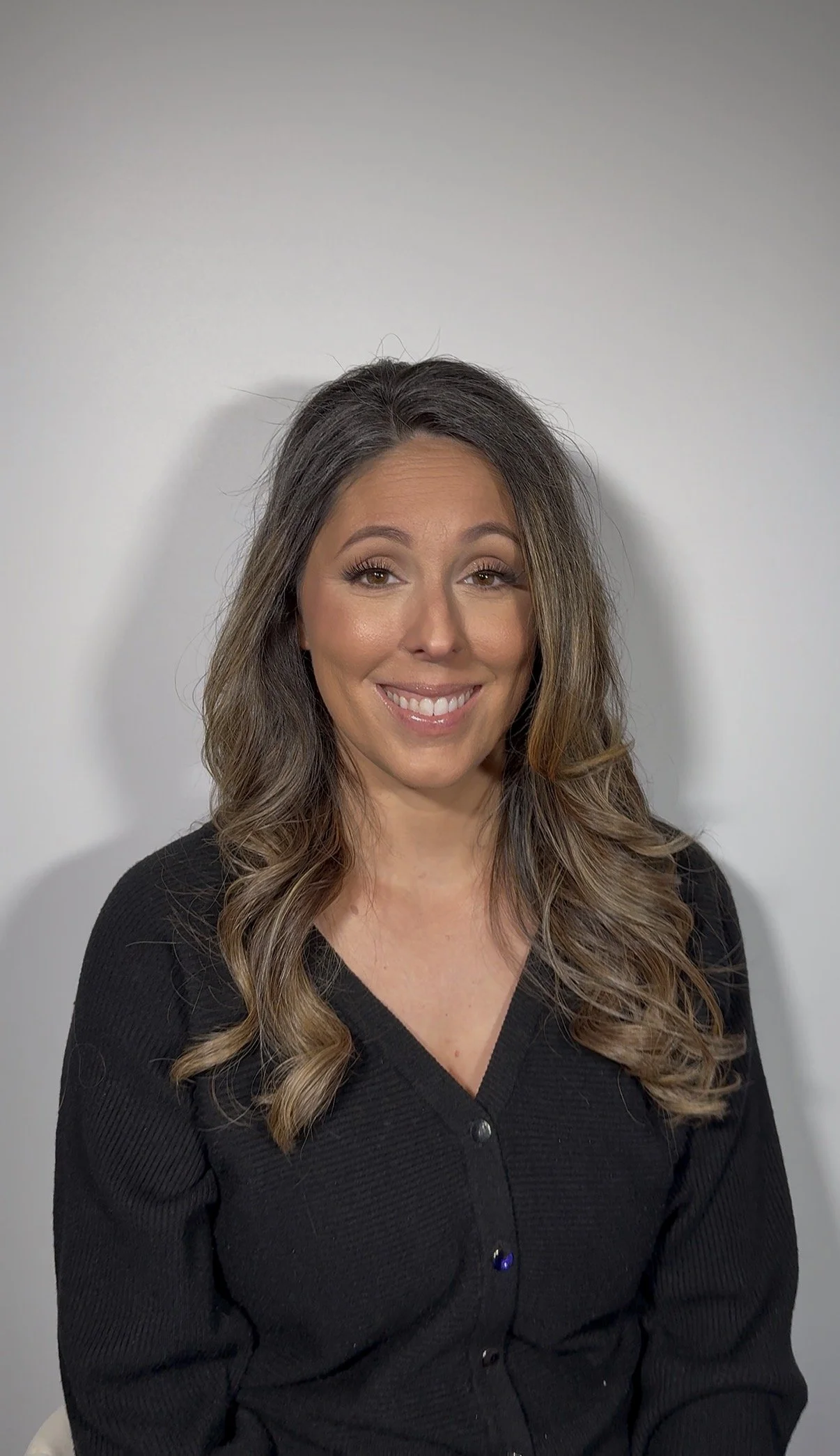 A woman with long, wavy brown hair and a warm smile, wearing a black ribbed cardigan, is sitting against a plain light gray background.