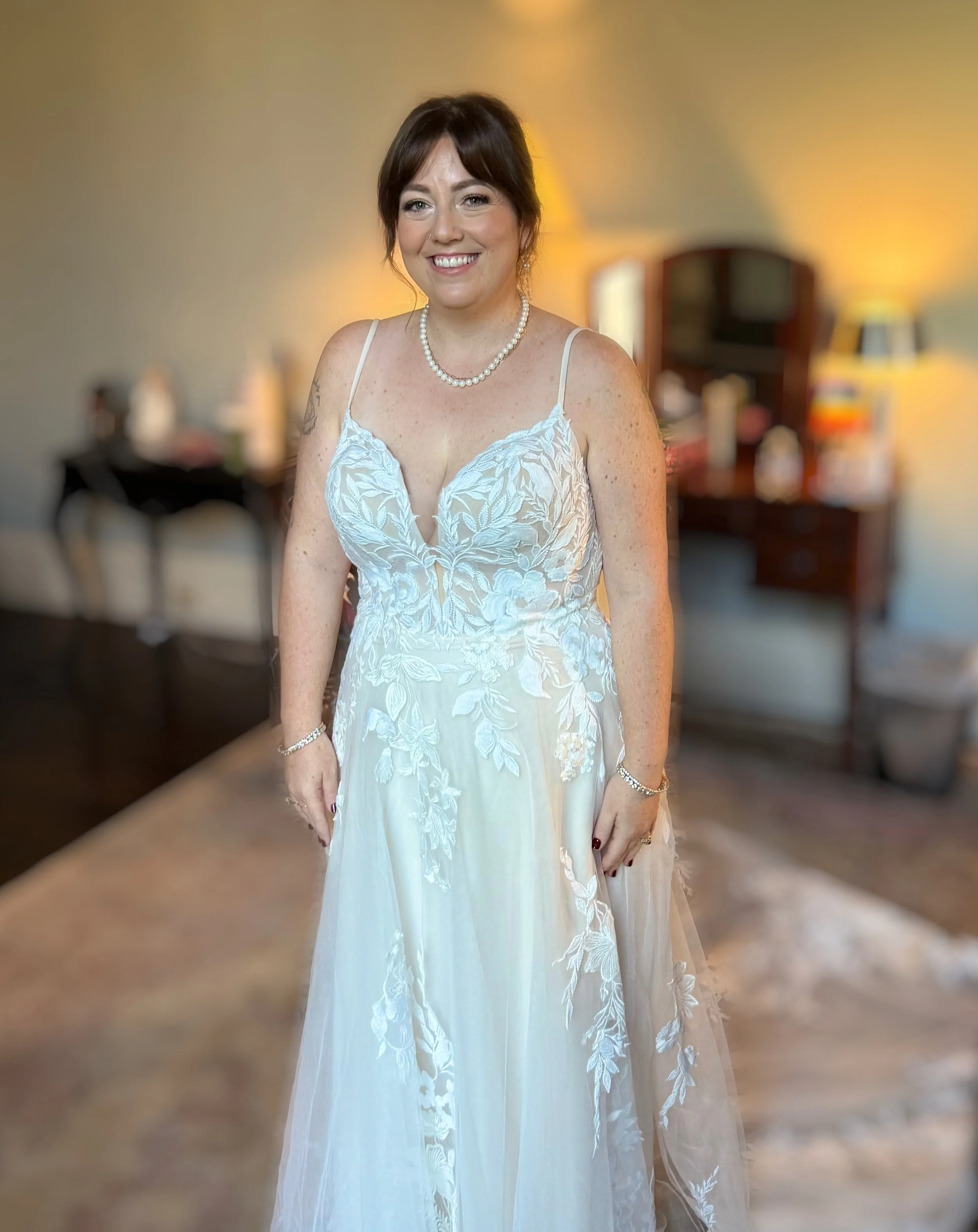 A woman with fair skin and dark brown hair styled in a loose updo, smiling in a room with warm lighting. She wears a white lace dress with spaghetti straps, a pearl necklace, a bracelet, and a ring, with a tattoo on her right arm. The background feat