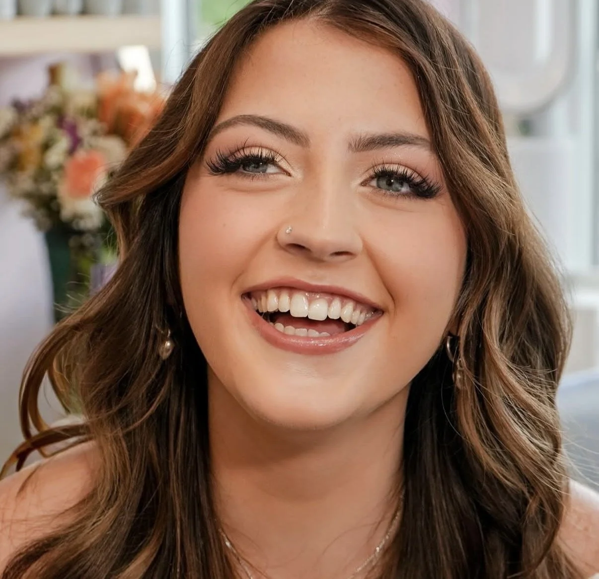 A young woman with long brown hair, bright blue eyes, and a nose piercing, smiling and showing off her teeth with a joyful expression, indoors with flowers in the background.