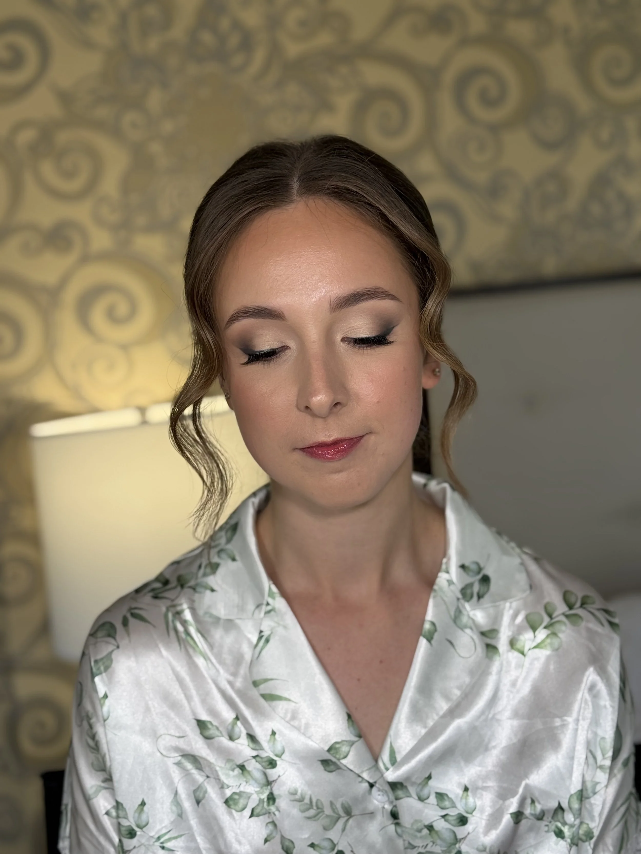 A woman with styled hair, makeup, and a satin robe with a green leaf pattern, sitting in a room with a gold and black patterned wall behind her.