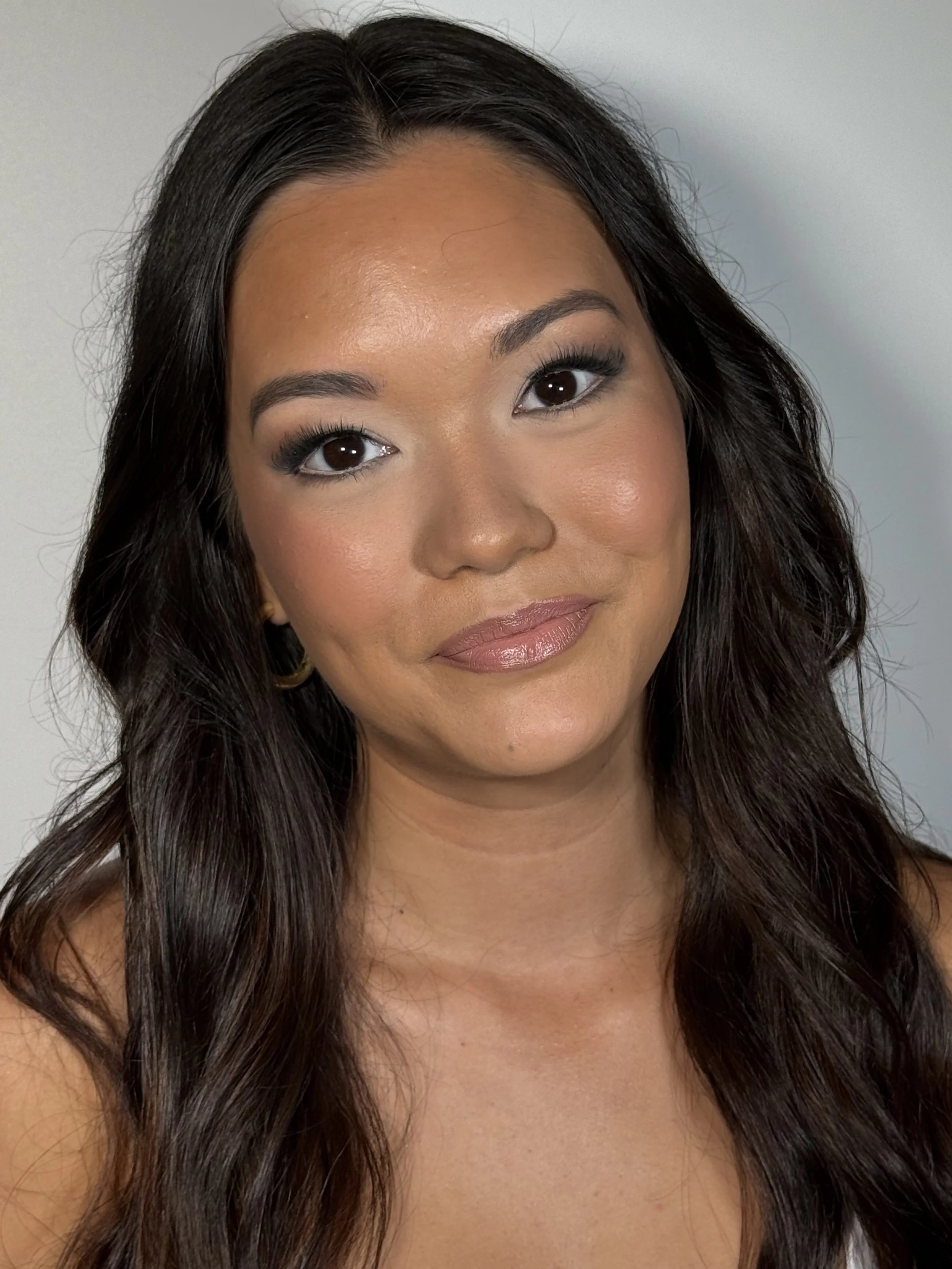 A close-up photo of a woman with long dark hair, smiling slightly, with makeup and earrings.