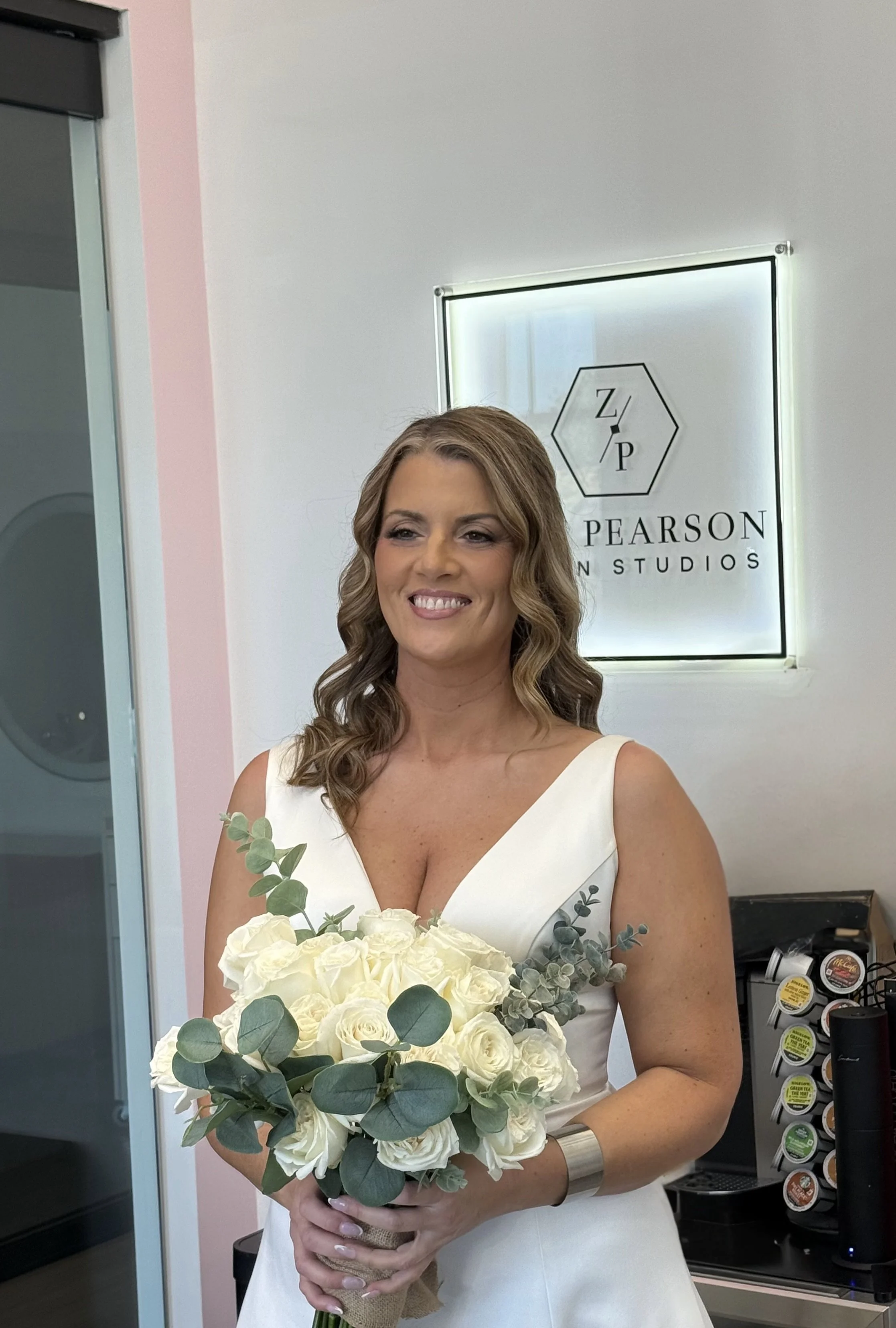 A smiling woman in a white dress holding a bouquet of white roses and eucalyptus in a room with a sign behind her that reads "Z/P Pearson Studio."