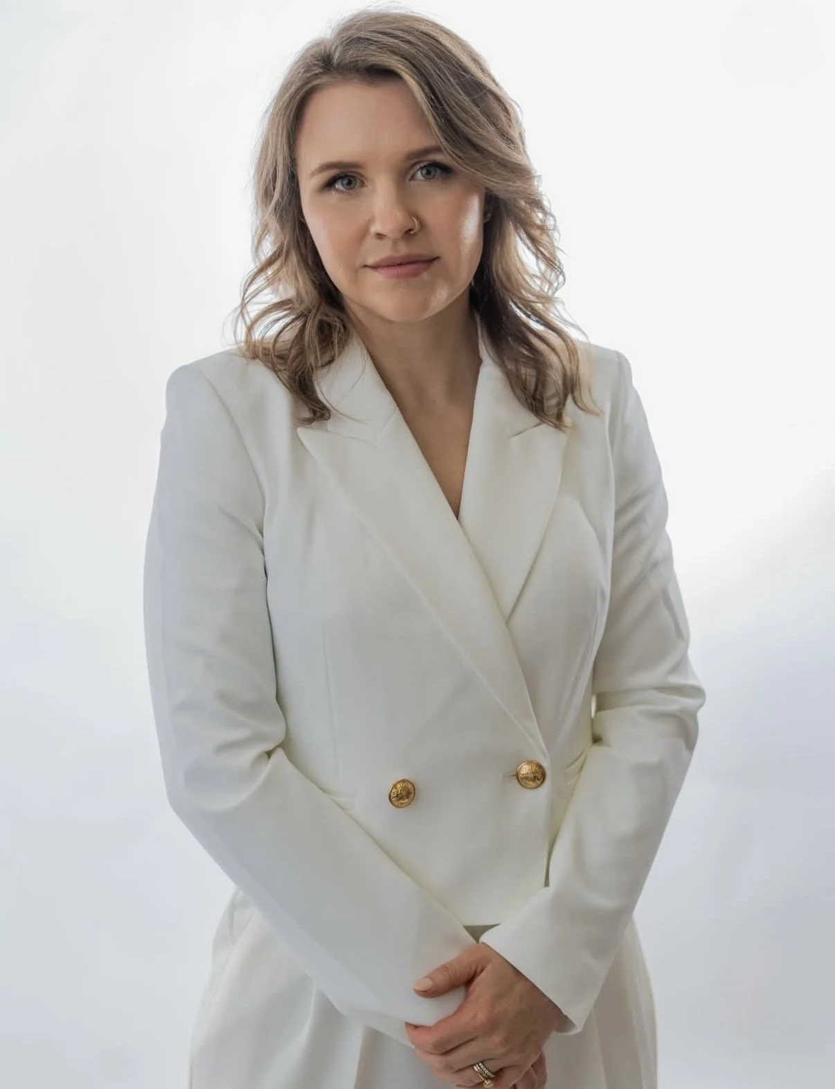 A woman with shoulder-length blond hair, wearing a white blazer with gold buttons, stands against a plain white background.