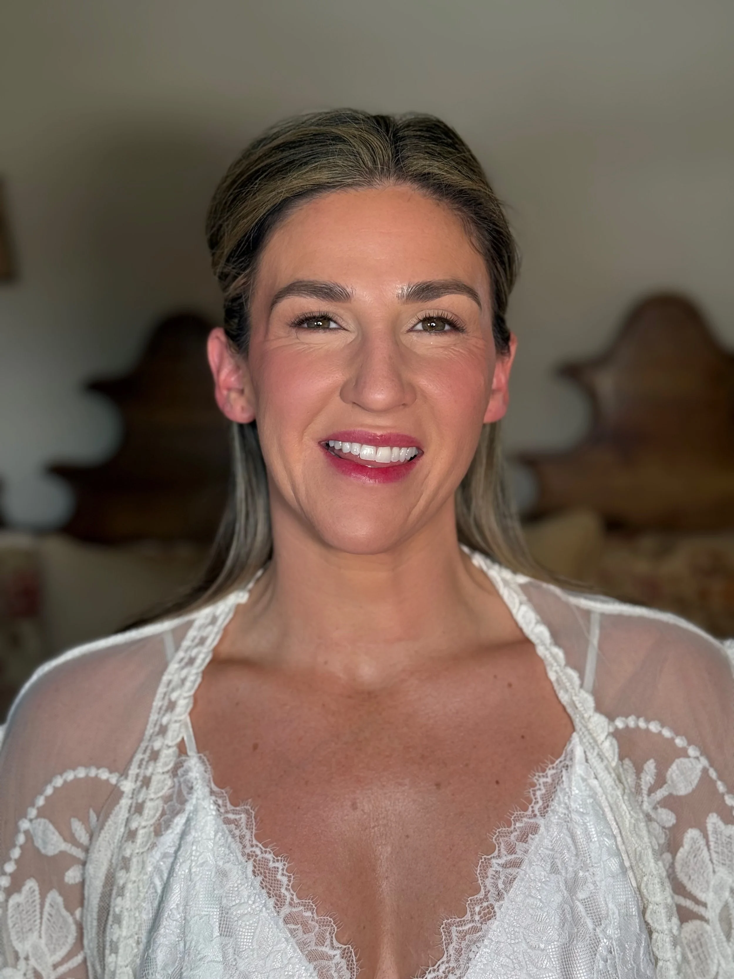 A woman with blonde hair styled back, wearing a white lace wedding dress with sheer sleeves, smiling at the camera, with a wooden headboard in the background.