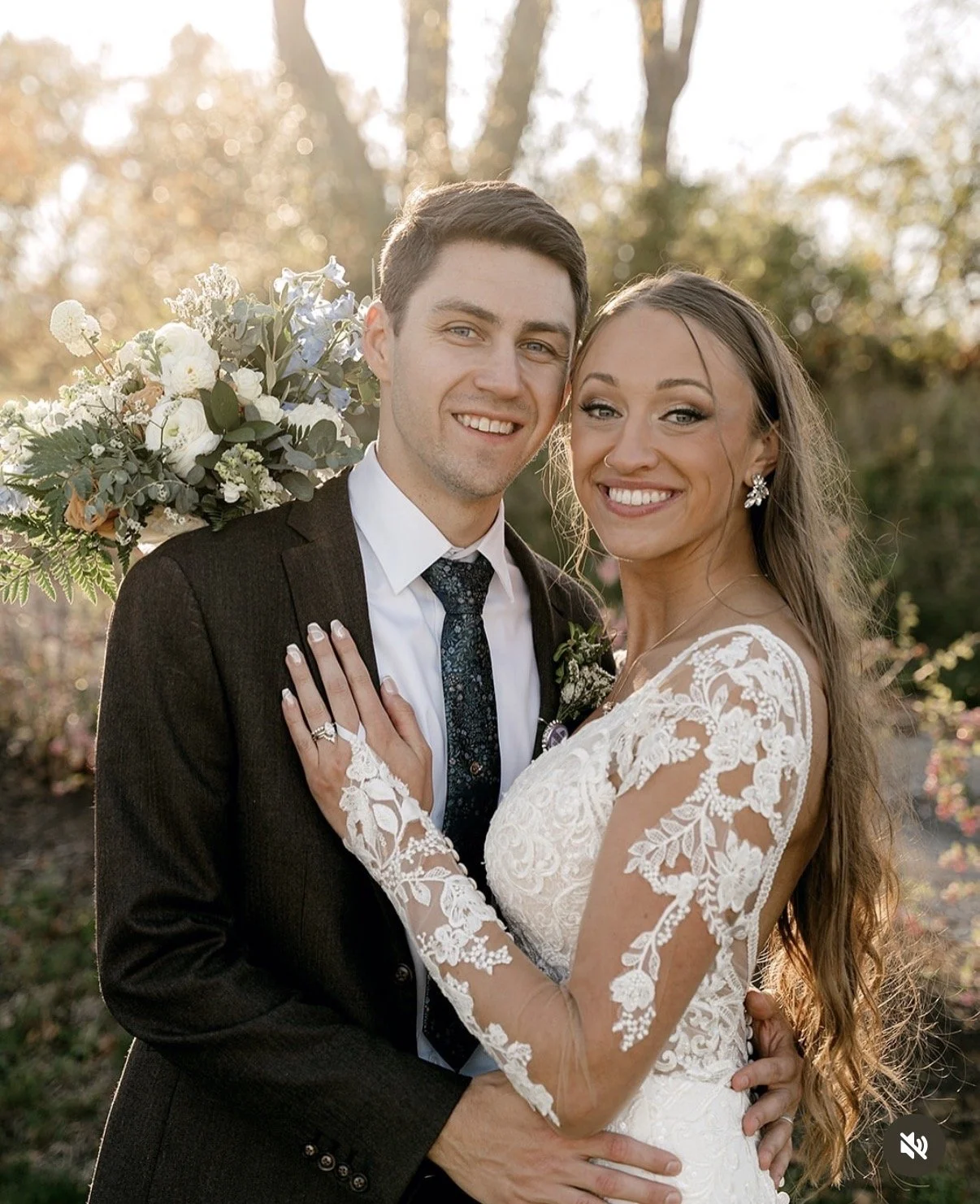 A newlywed couple smiling and posing outdoors in a garden setting during sunset. The groom is wearing a dark suit with a tie, and the bride is in a lace wedding dress with long sleeves, holding a bouquet of flowers behind her back.