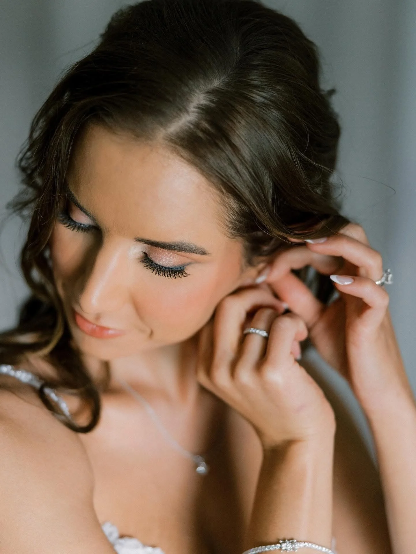 Close-up of a woman with brown hair styled in loose curls, wearing jewelry, gently touching her ear, with eyes closed and a serene expression.