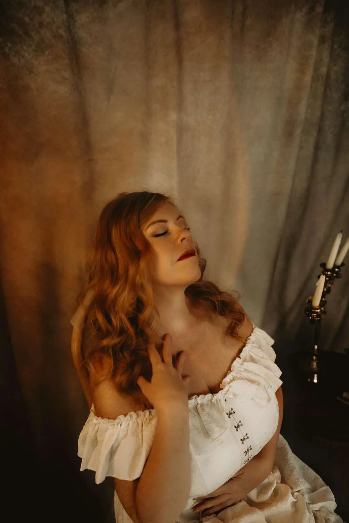 A woman with long, wavy red hair, wearing a white dress with embroidery, is resting her hand on her chest with her eyes closed, in a dimly lit room with a dark background and a candelabrum with candles.