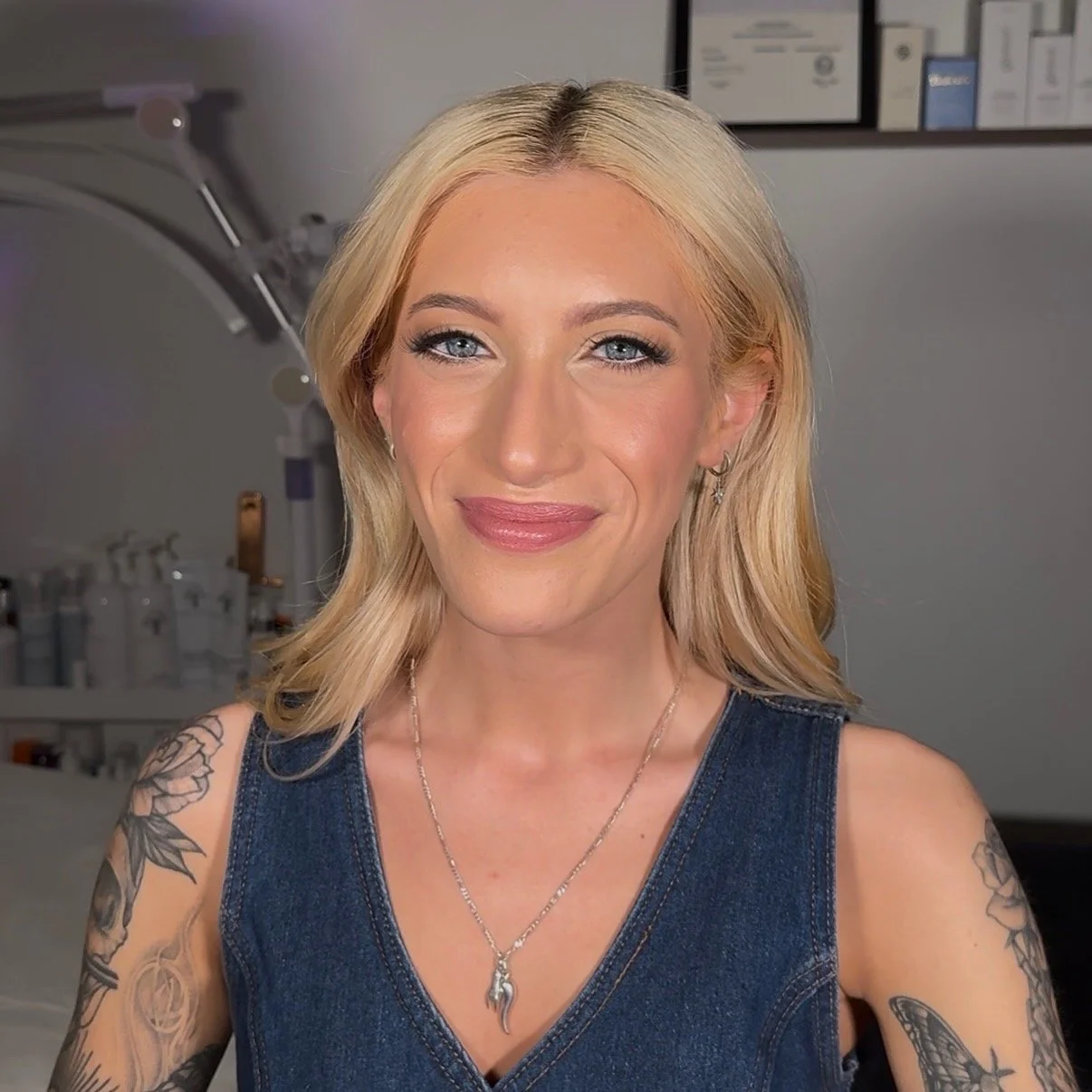 A smiling woman with blonde hair and tattoos on her arms, wearing a sleeveless denim top and a silver necklace with a pendant, in a medical or beauty clinic setting.