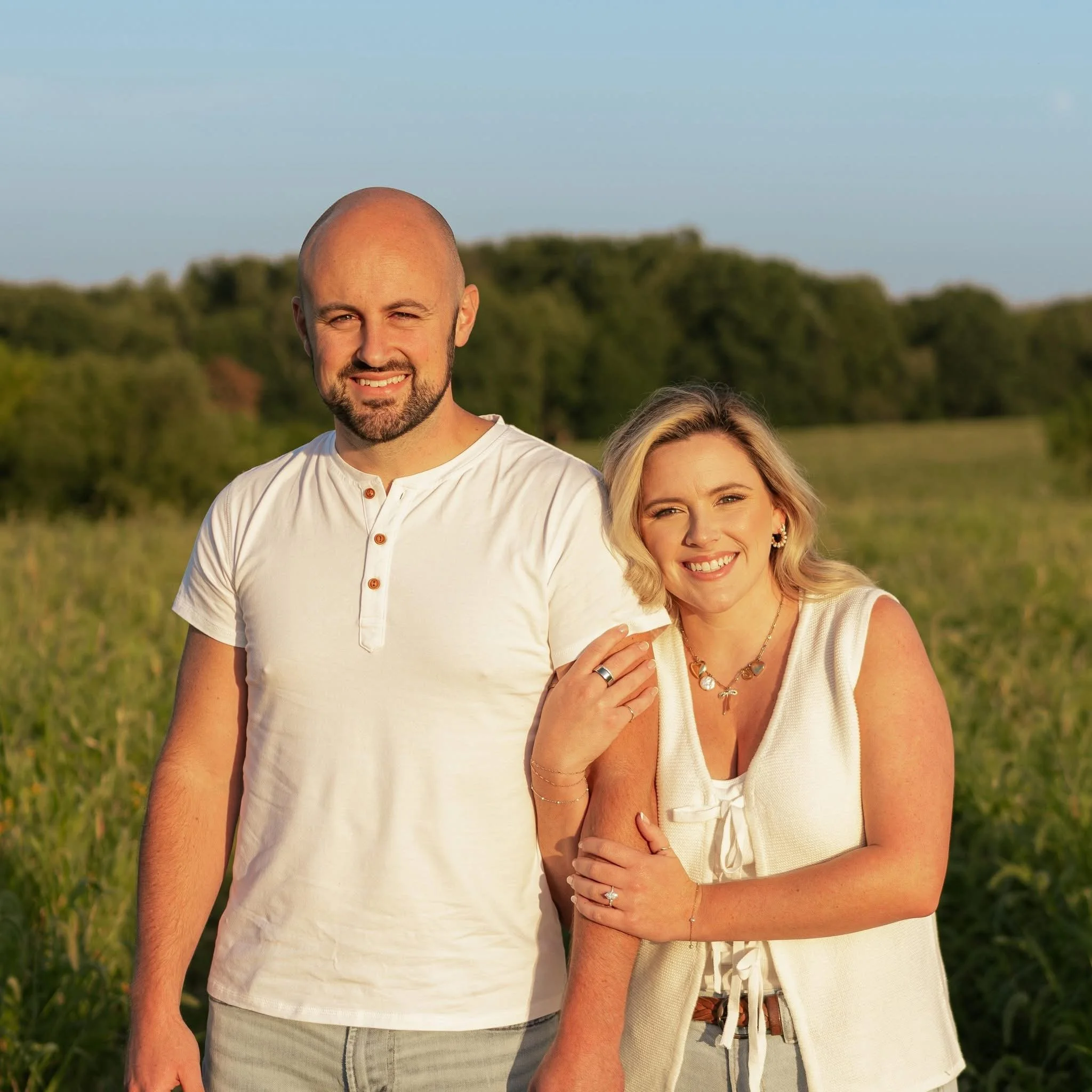 Close-up of a smiling man with a beard and woman standing outdoors in a field during sunset, with a background of trees and a clear sky.