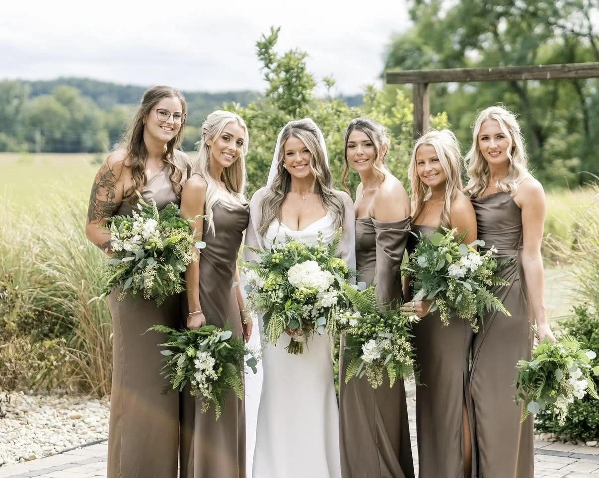 A bride and five bridesmaids standing outdoors with greenery in the background, holding bouquets of white and green flowers, dressed in taupe gowns.