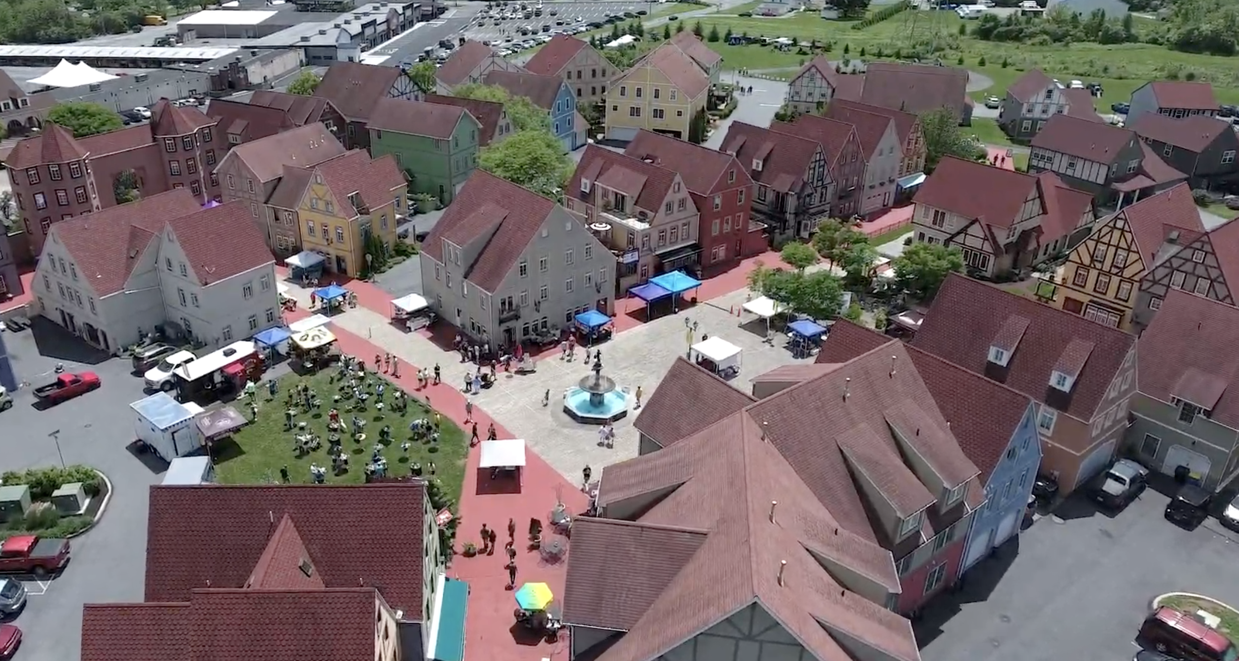 Aerial view of a colorful town square with a fountain, surrounded by pastel houses, tents, and people attending an outdoor event on a sunny day.