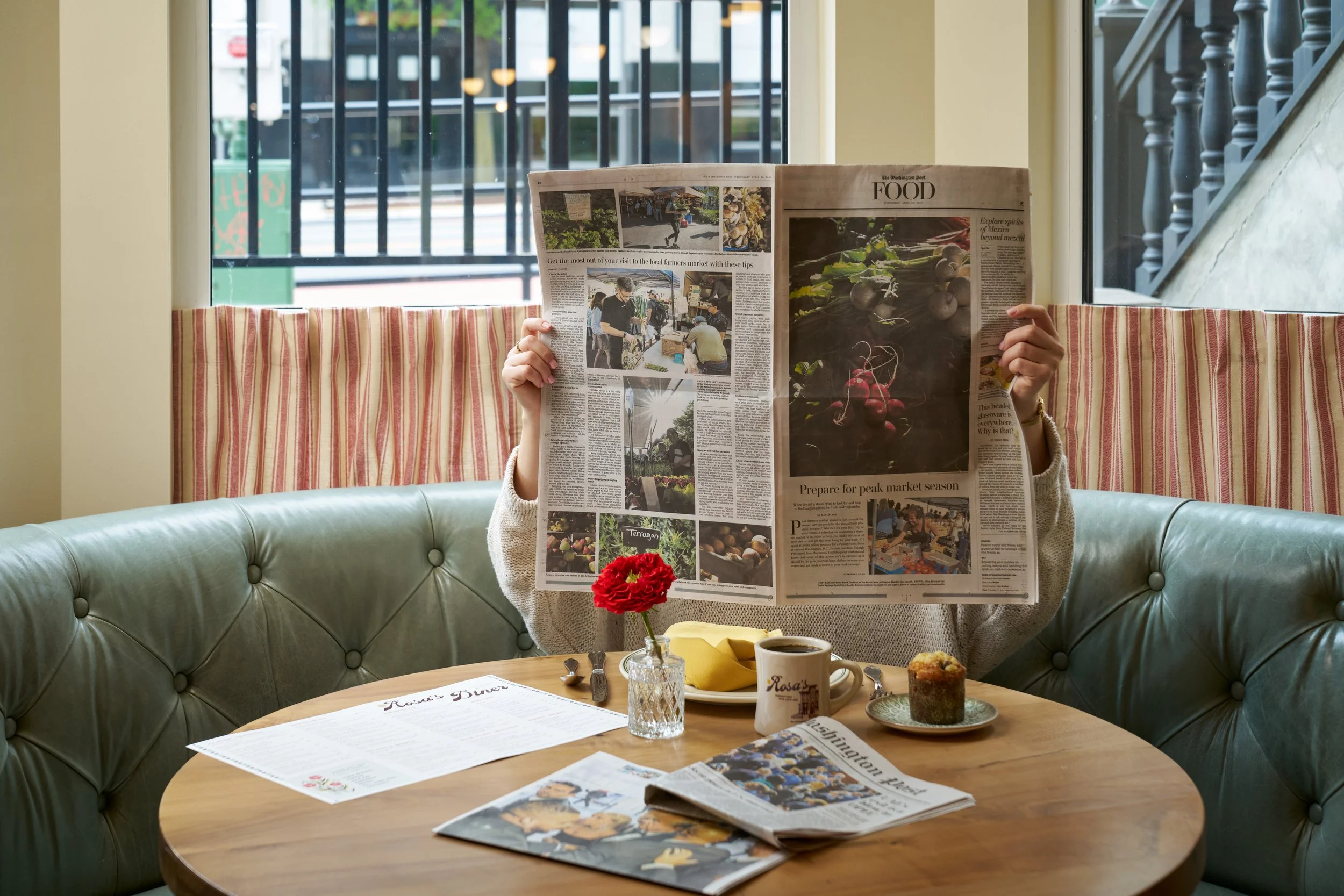 Person sitting in a booth at a restaurant, reading a newspaper with a green leather table and a small flower in a glass cup. Breakfast items on the table, including a muffin, a cup of coffee, and printed menus. Large windows in the background showing outdoor scenery.