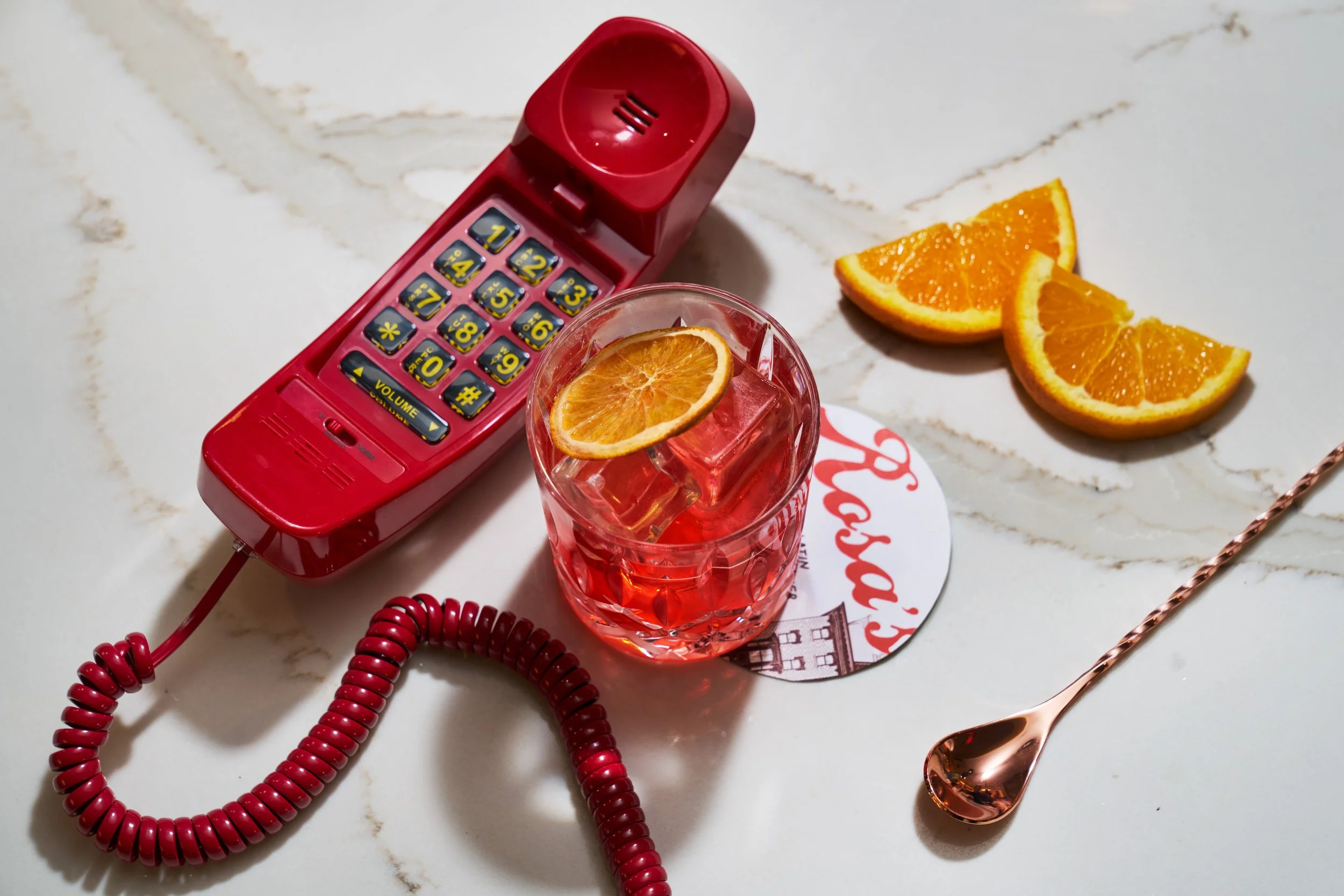 A vintage red telephone, a glass of iced tea with a slice of orange, and three orange slices on a white marble surface.