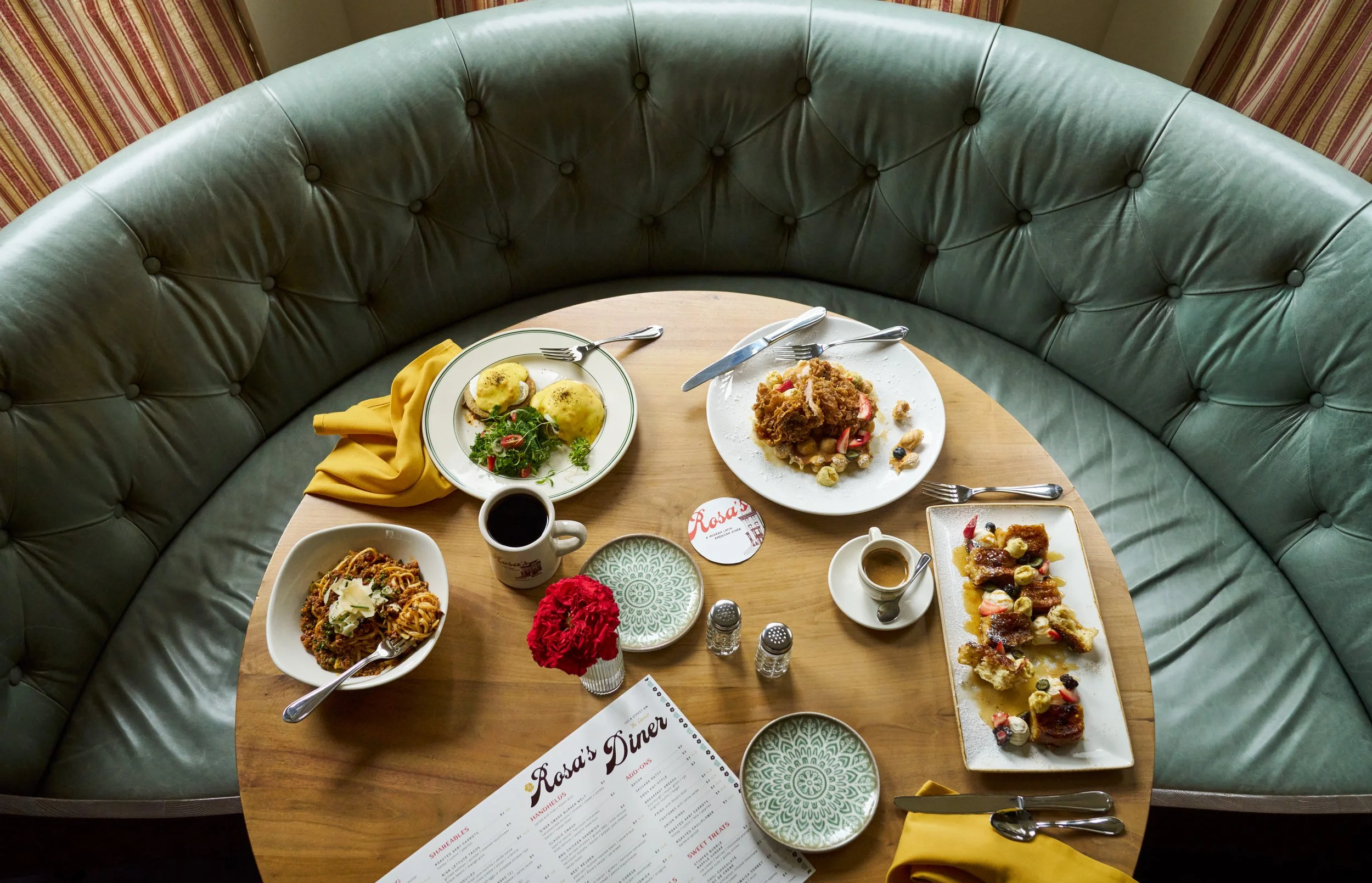 A table set with various breakfast dishes including eggs Benedict, pasta, and fruit, surrounded by a green satin banquette and other chairs in a cozy cafe setting.