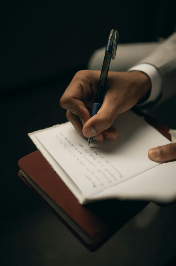 Person writing in a notebook with a pen, wearing a suit, with additional notebook underneath.
