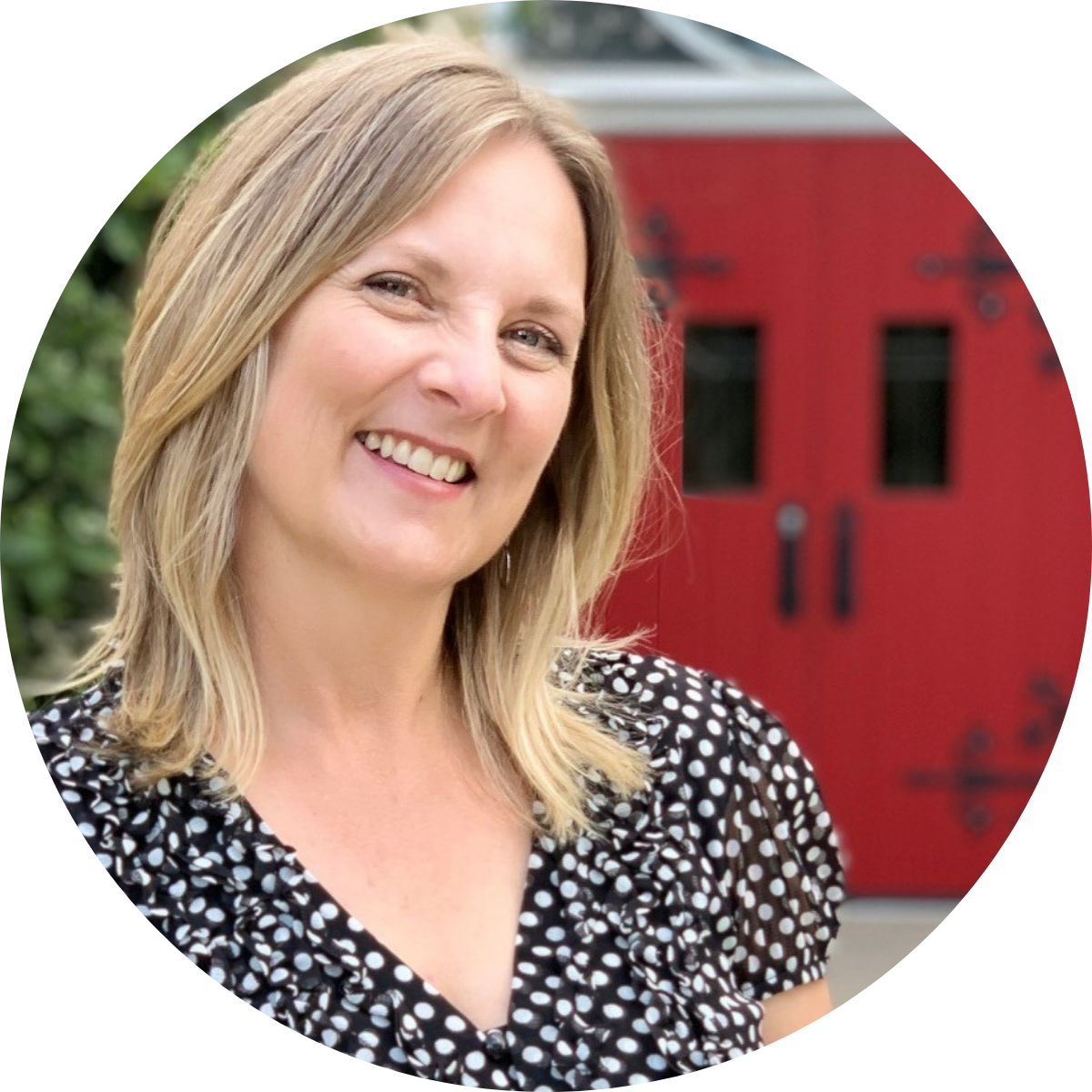 A smiling woman with blonde hair wearing a black and white polka dot blouse standing outdoors with a red building in the background.