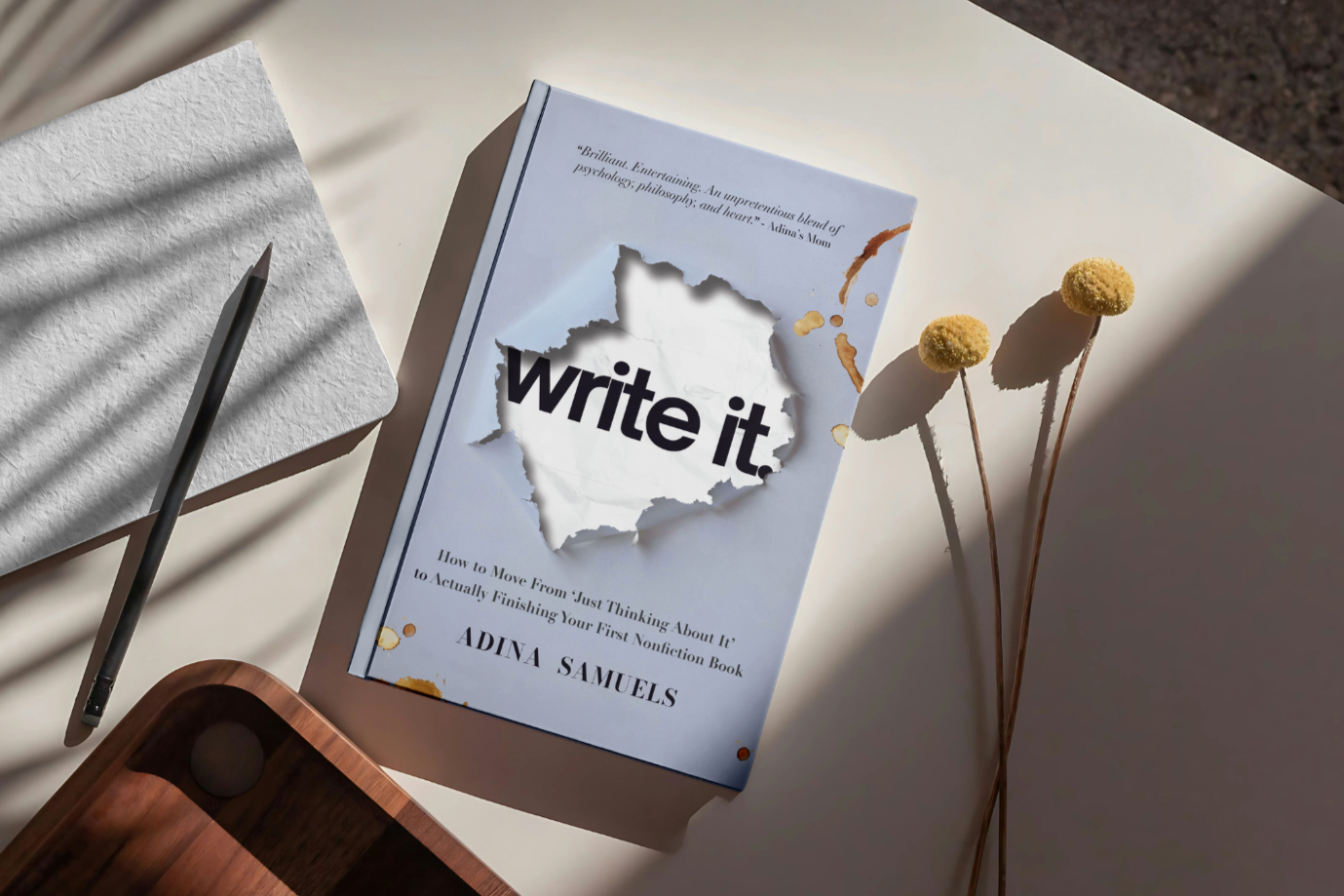 Book titled 'write it' on a white desk, with two dried yellow flowers and a gray pencil nearby, natural light casting shadows.