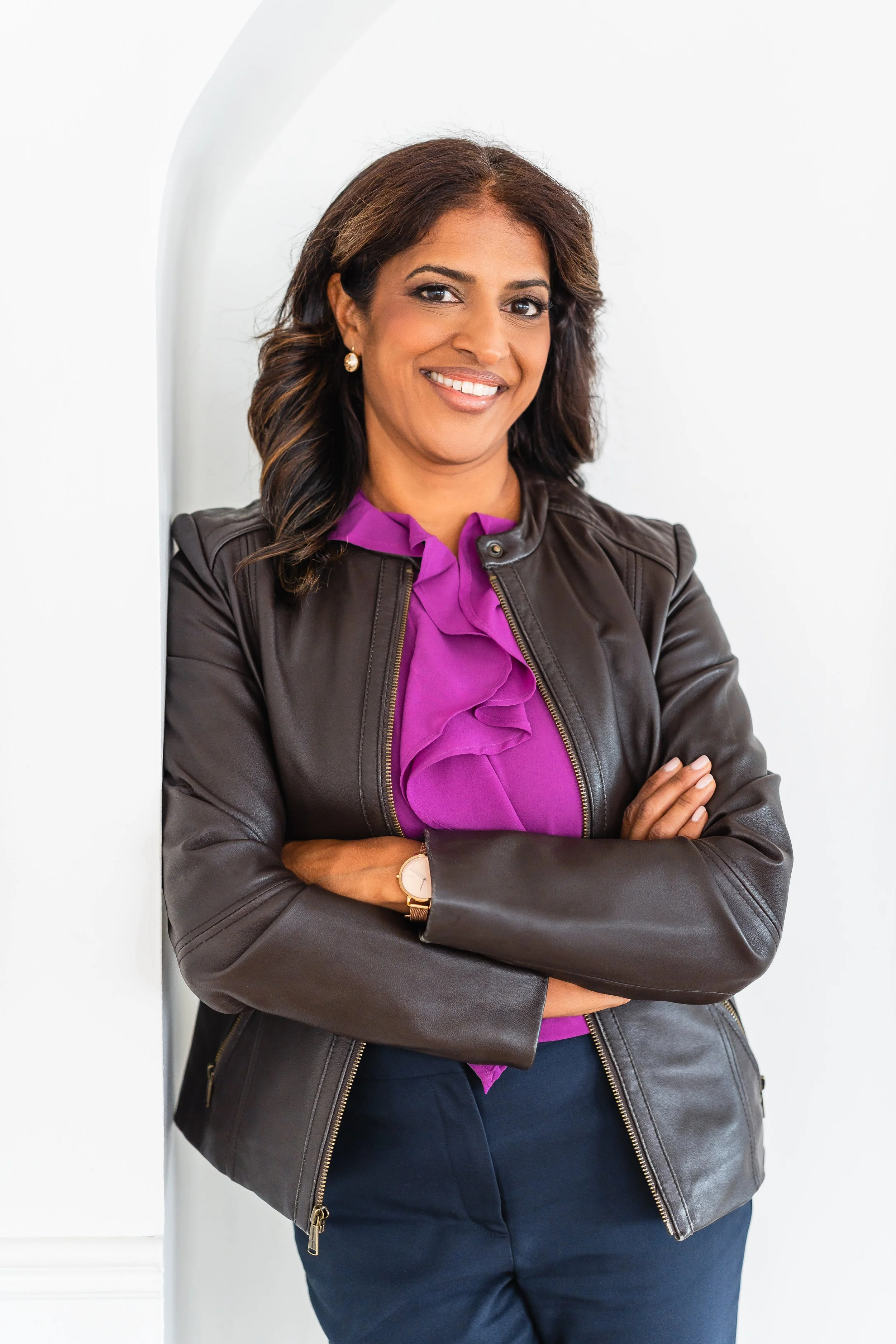 A woman with medium skin tone, shoulder-length brown hair, wearing earrings, a black leather jacket over a purple blouse, and dark pants, standing with arms crossed in front of a white wall.