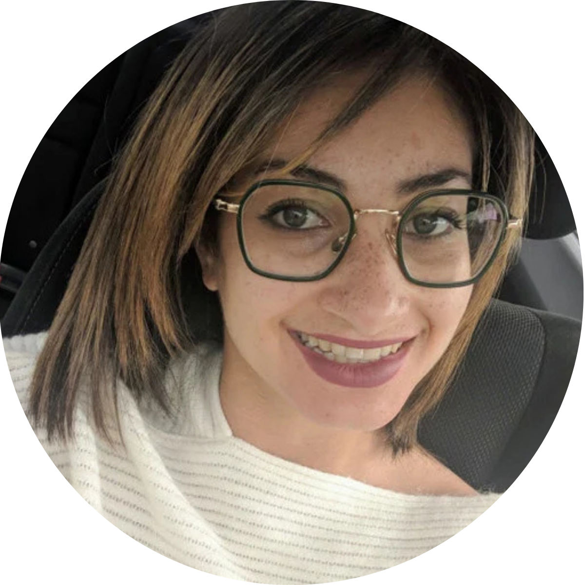 A young woman with shoulder-length brown hair, glasses, and freckles, smiling at the camera inside a vehicle.