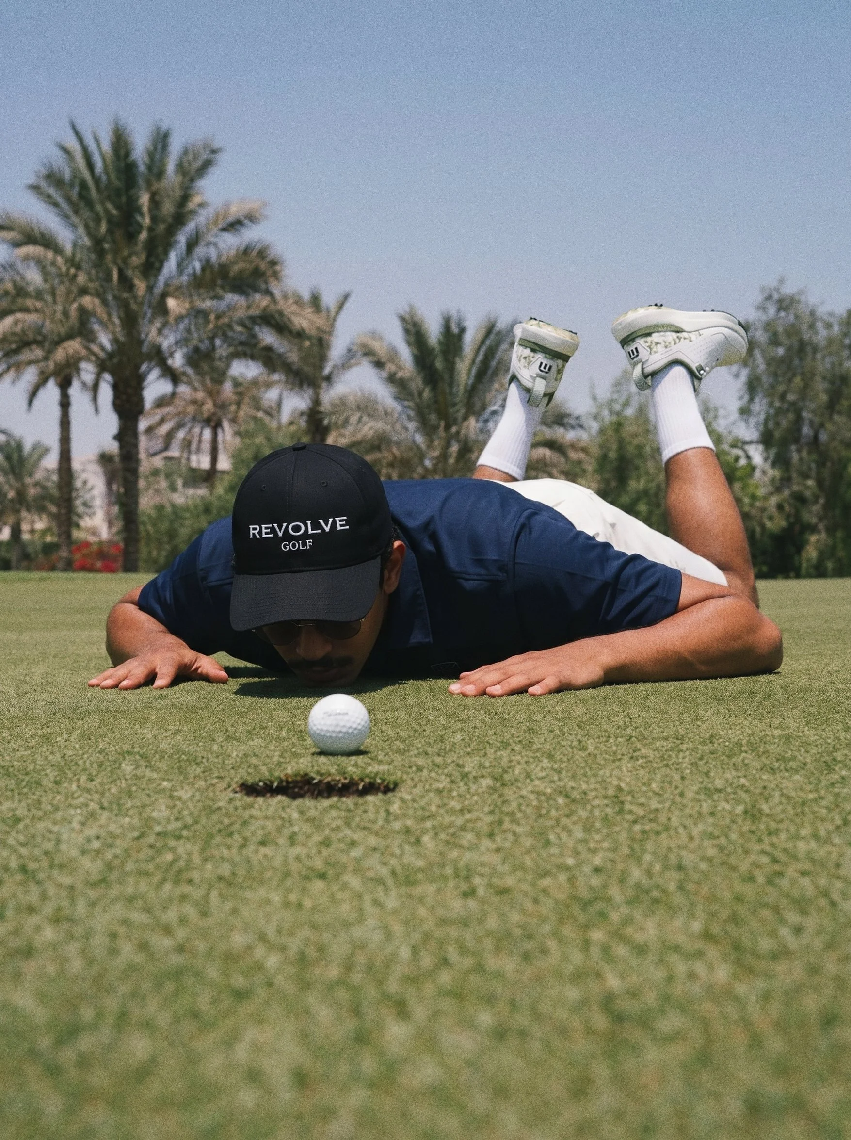 A person lying face down on a golf course putting green, with their legs raised behind them. The person is wearing a black cap with the words 'REVOLVE GOLF' and white golf shoes. A golf ball is positioned near the person's hand and hole.