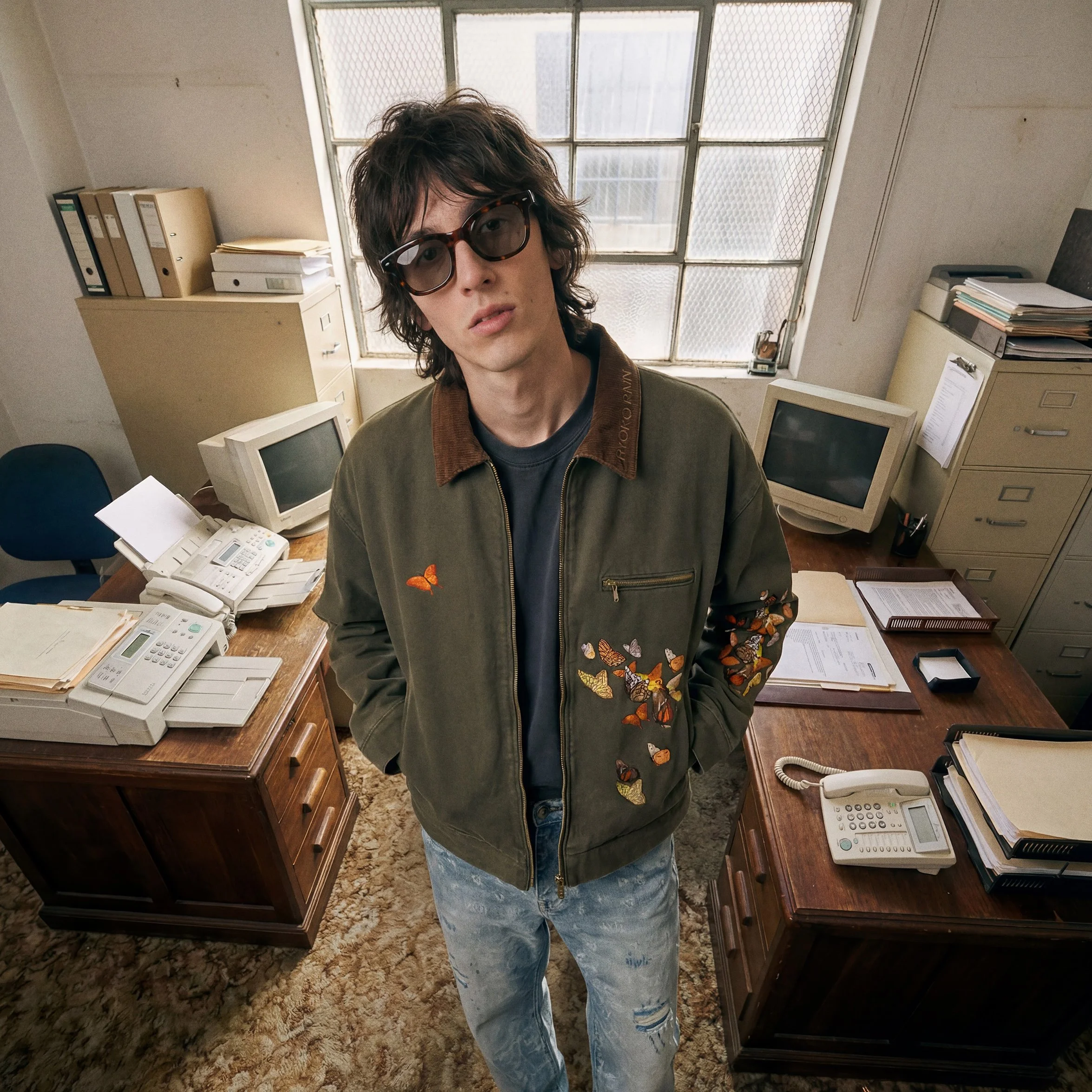 A person with wavy brown hair wearing sunglasses, a green jacket with butterfly patches, and ripped jeans. They are standing in an office with old computers, filing cabinets, and paperwork.