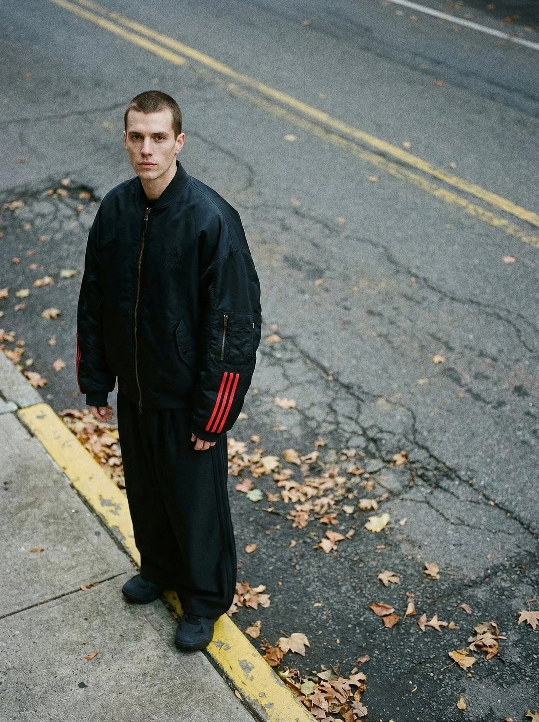 A young man standing on a street curb with yellow double lines, wearing a black Adidas jacket and black pants, surrounded by fallen autumn leaves.