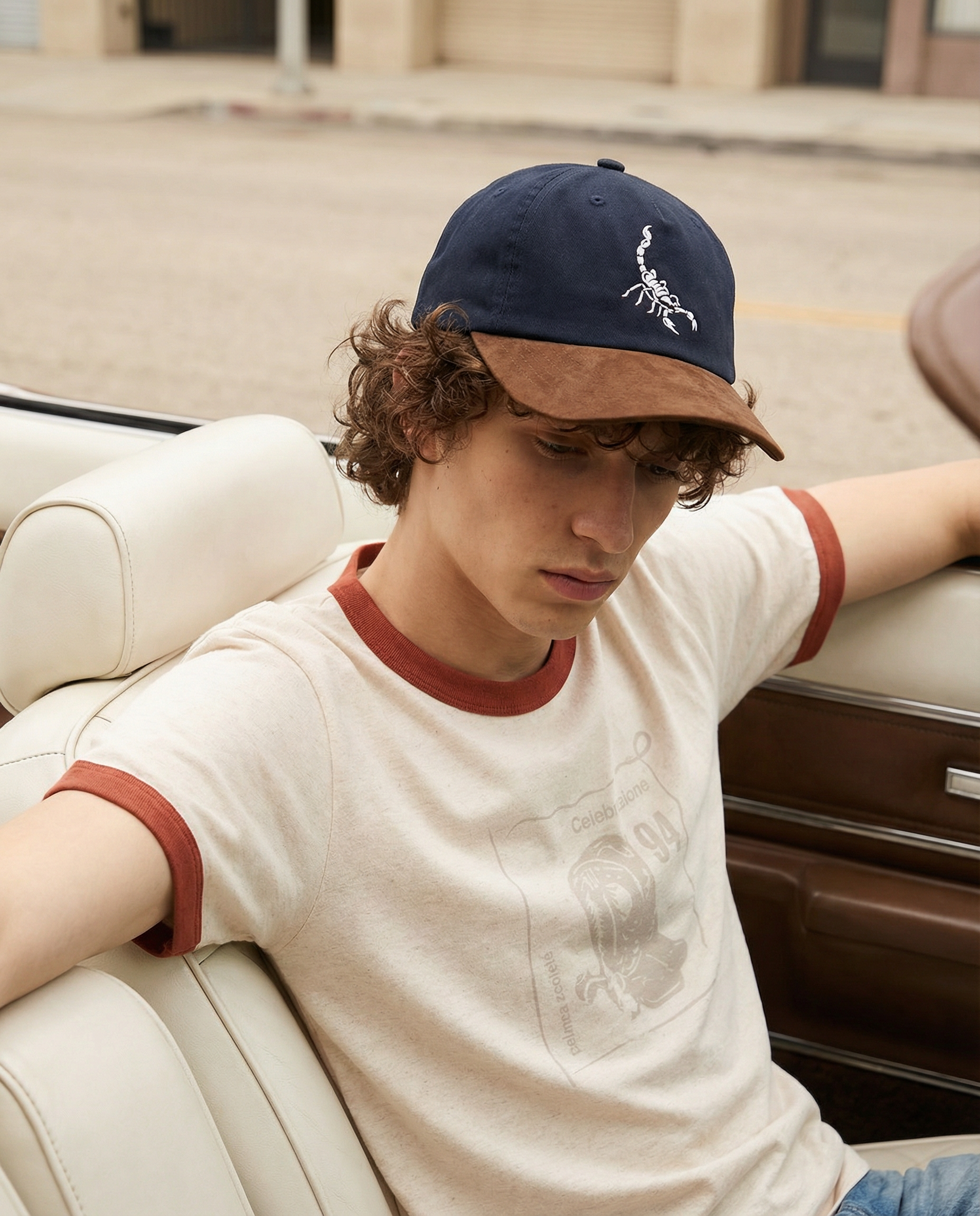 A young person with curly hair is sitting on a white couch, wearing a navy blue baseball cap with a brown brim and a white design of a scorpion. They are looking down and resting their arm on the back of the couch. The background shows a sandy area a