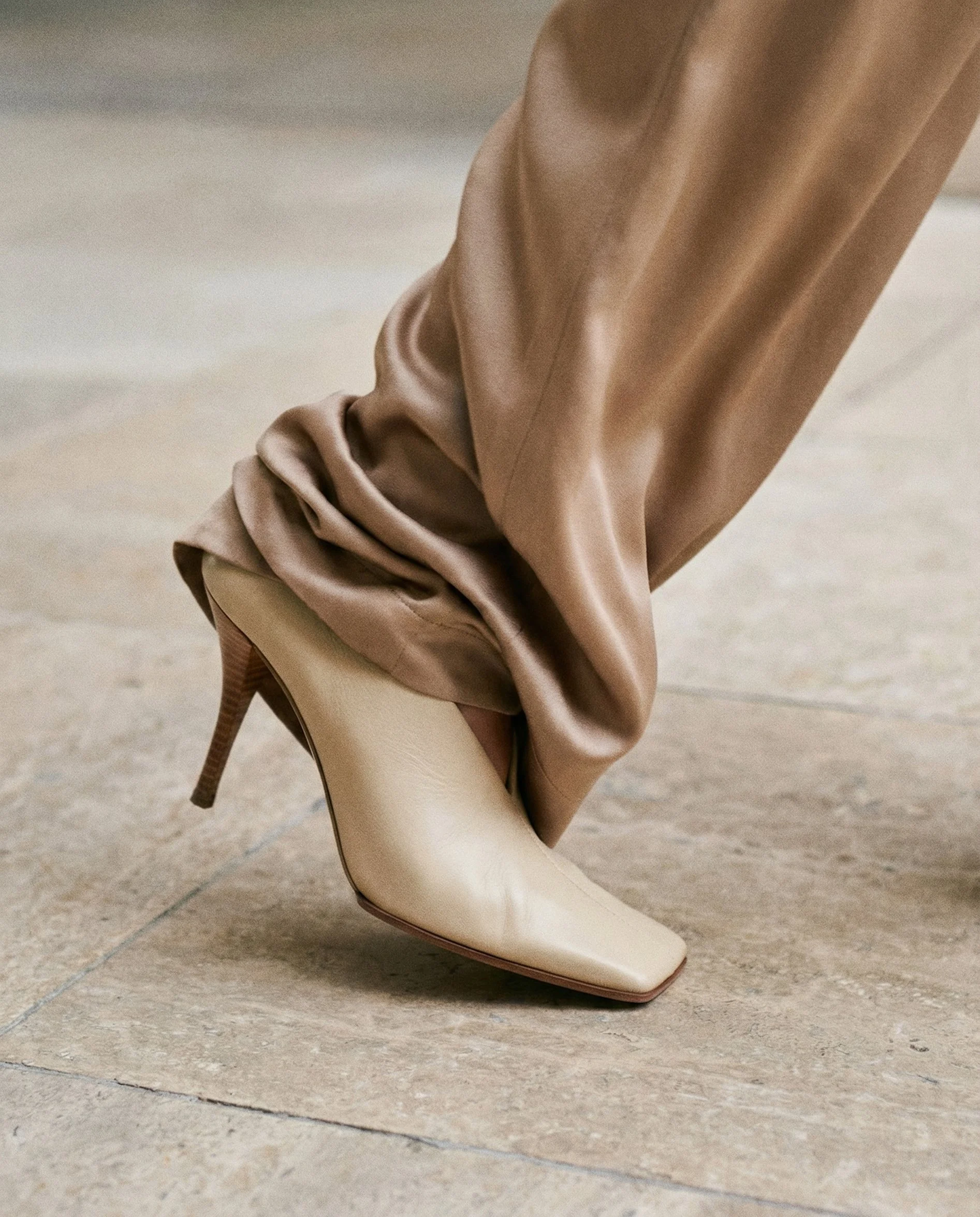 Close-up of a beige high-heeled shoe with a pointed toe, placed on a tiled floor, with a brown fabric draped over the shoe.