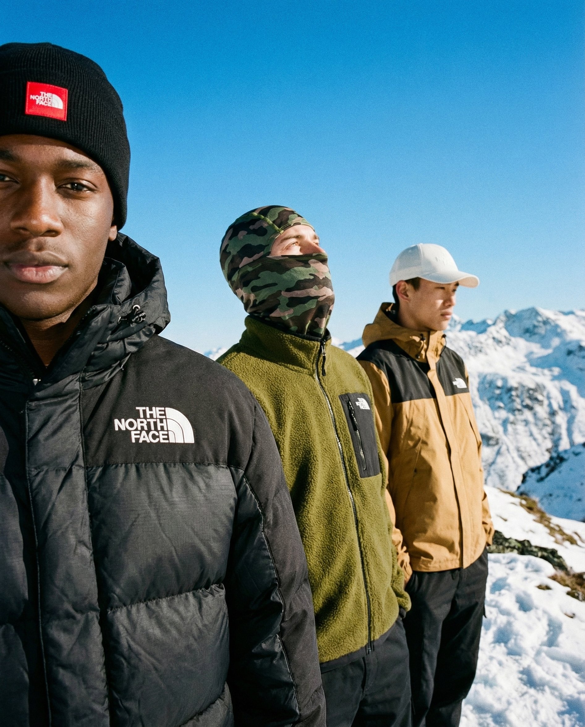 Four men standing outdoors on snow-covered mountains wearing winter jackets and hats, with a clear blue sky overhead.  AI fashion editorial image.Polo Ralph Laurent.