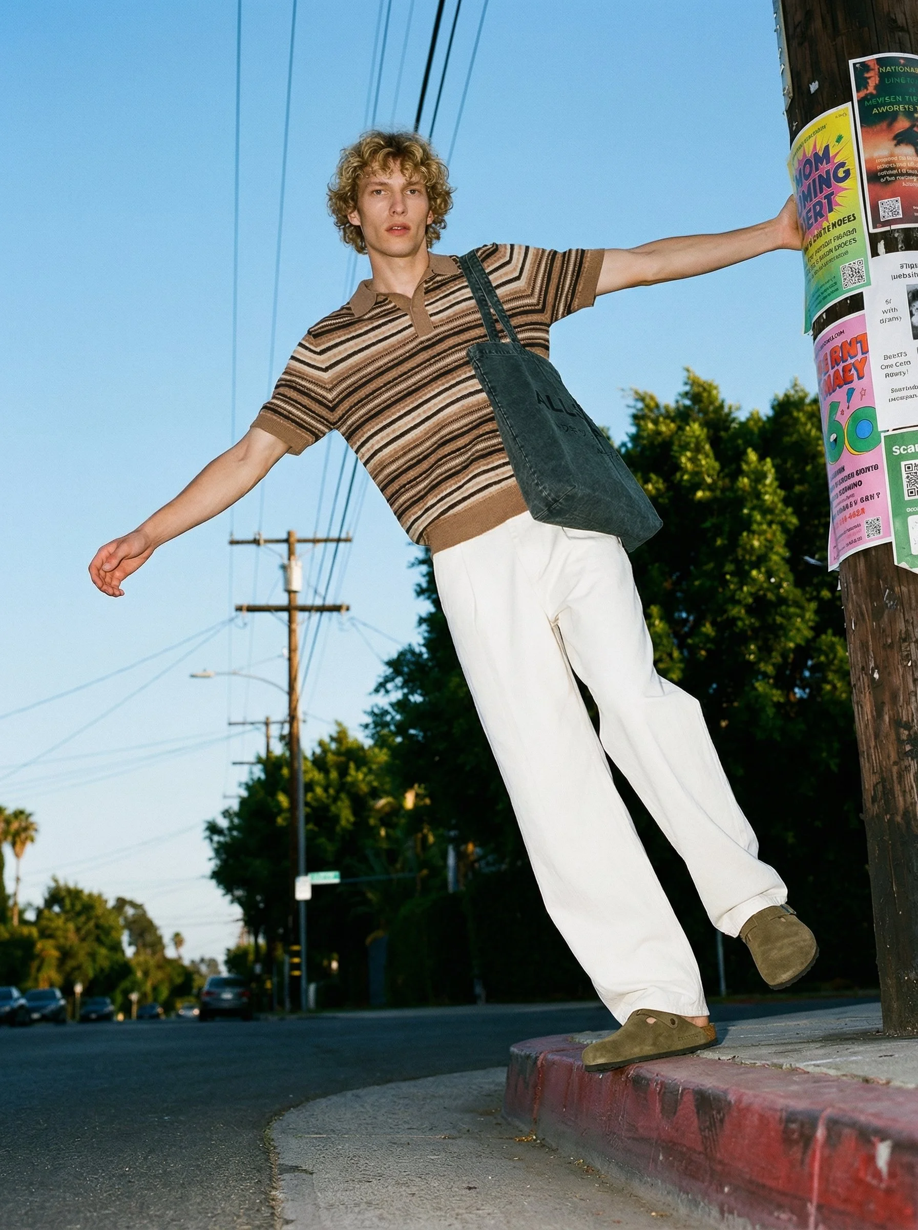 Young man with curly hair wearing a striped shirt, white pants, and green shoes standing on a curb with arms extended outward, next to a utility pole covered with colorful flyers, in a suburban street scene during daytime.