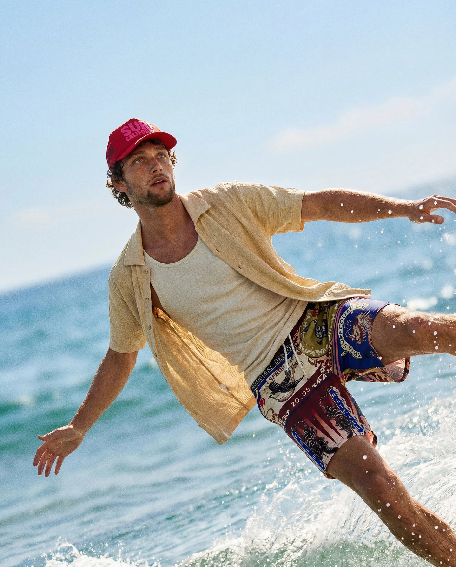 A man wearing a red cap, yellow shirt, and patterned shorts is surfing in the ocean, with blue sky and water in the background. AI fashion editorial image . AI men fashion.