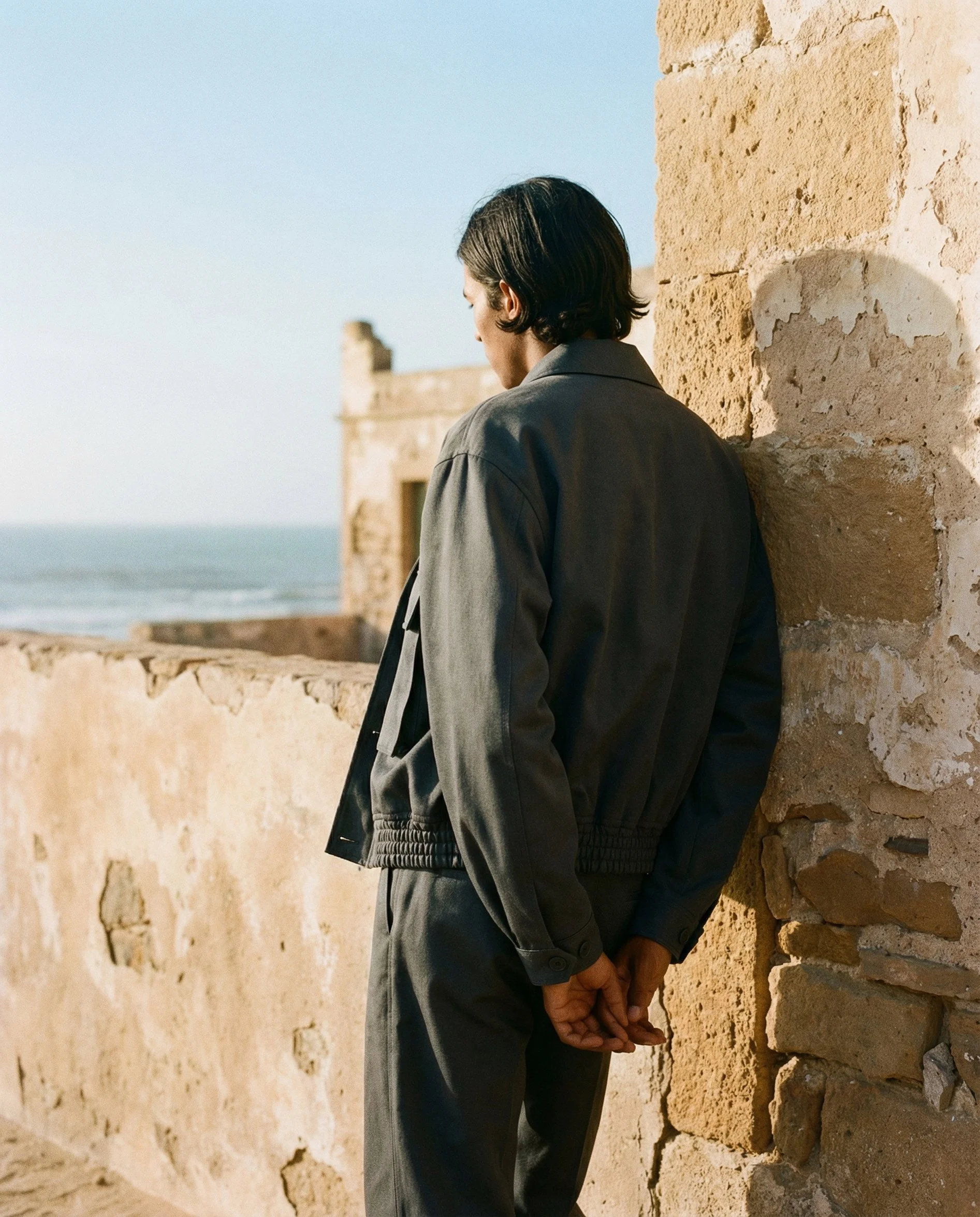 A man with dark hair stands with his back to a stone wall, facing the ocean on a beach, with a distant building visible.