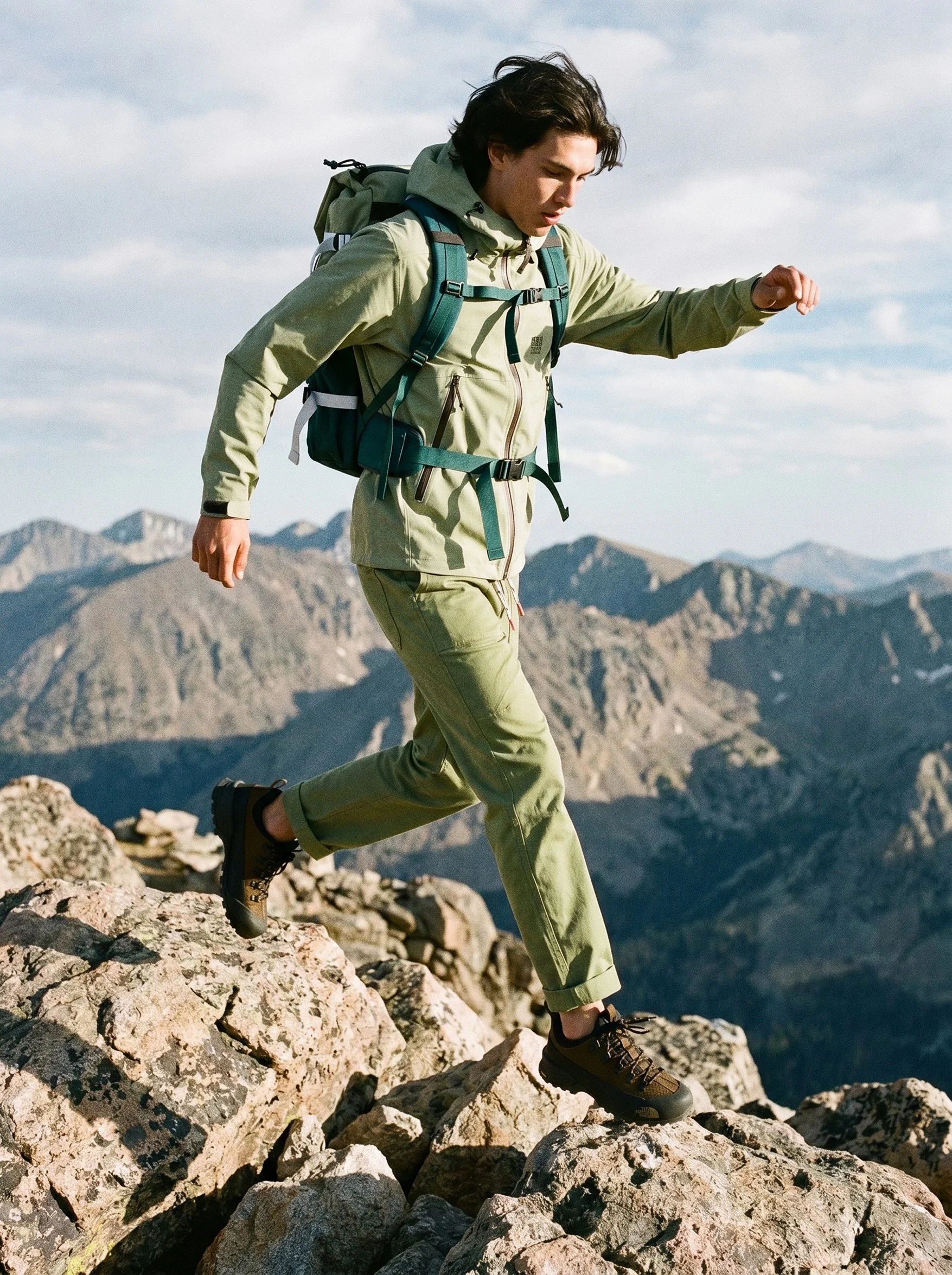 A young man hiking on rocky terrain in a mountainous area during daytime, wearing a green outdoor outfit, a backpack, and hiking boots.