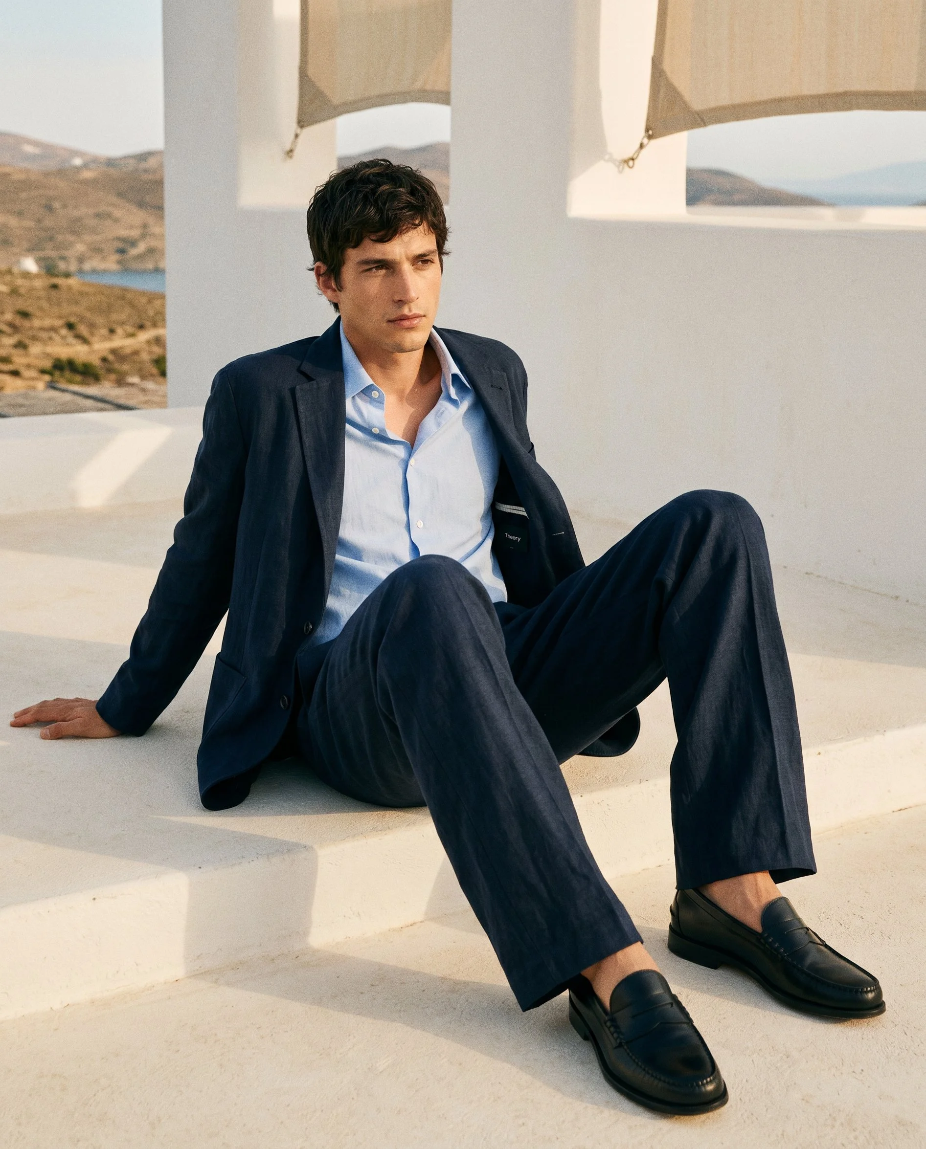 A young man in a suit sitting on white stairs outdoors with a landscape of hills and water in the background, looking contemplative.