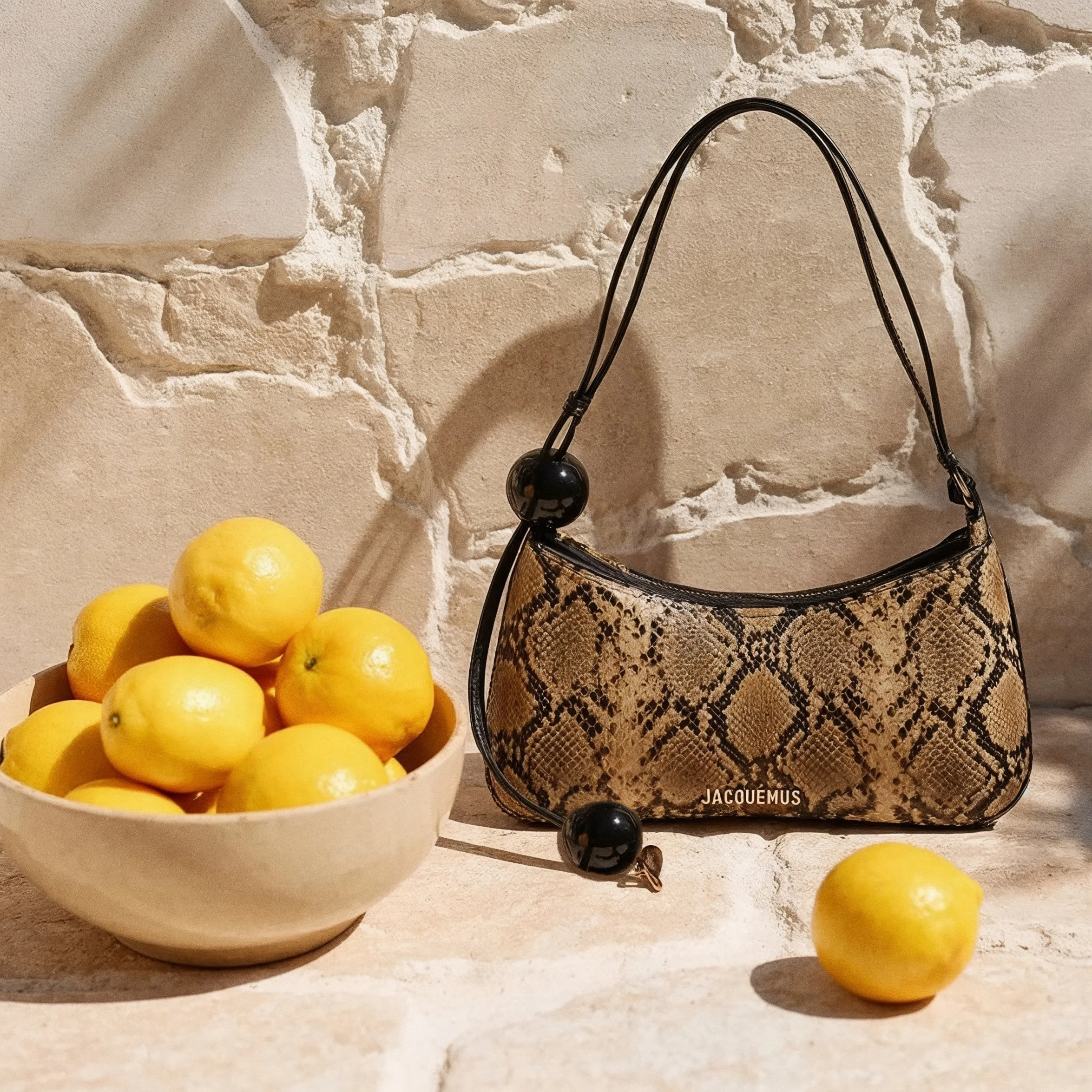 A white bowl filled with yellow lemons placed on a beige stone countertop. Behind the bowl, there is a textured white stone wall and a snakeskin-patterned purse with black straps hanging from it AI fashion editorial.