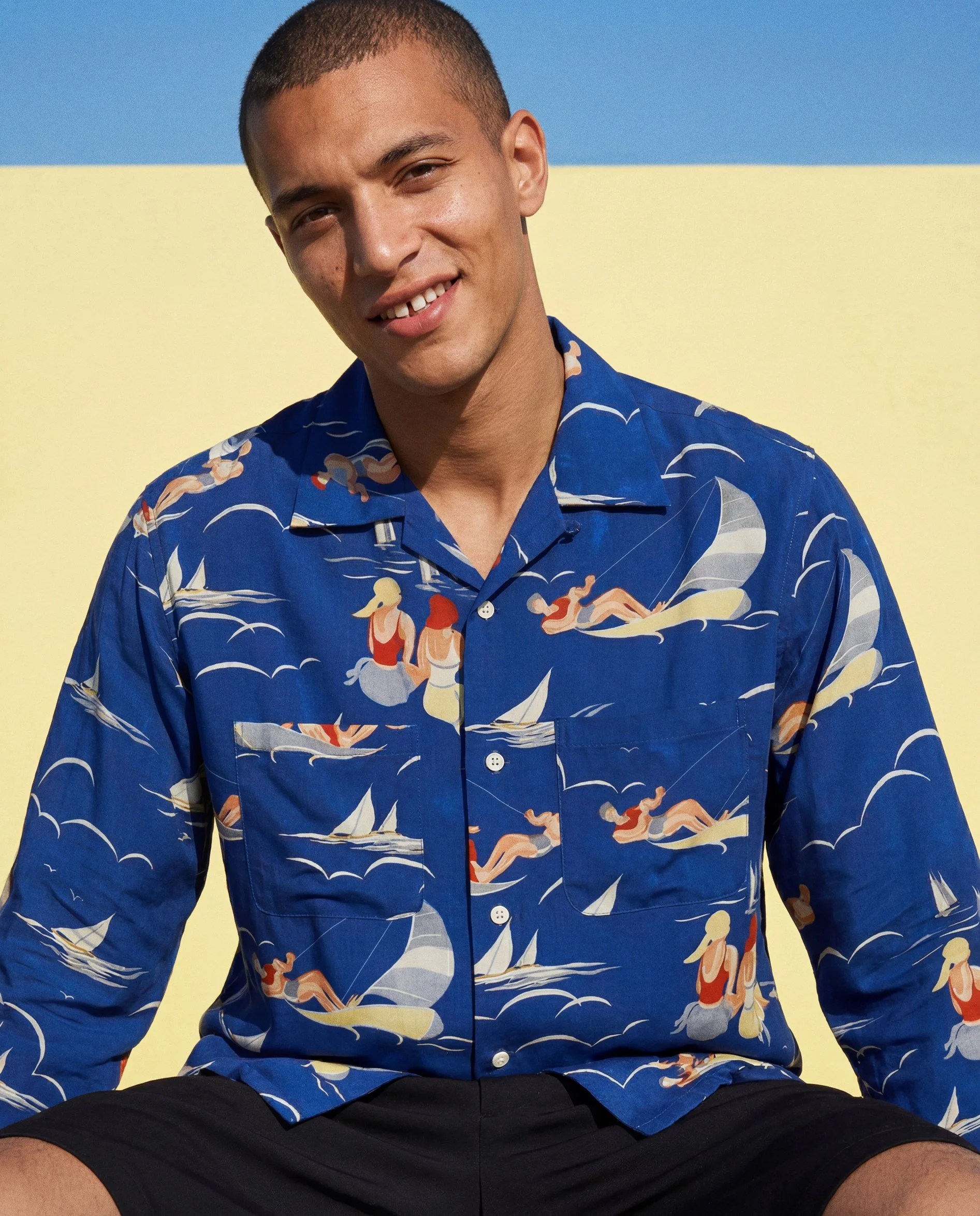 A young man with short hair, smiling and wearing a blue Hawaiian shirt with a pattern of waves and figures, sitting against a yellow and blue background.  AI fashion editorial.