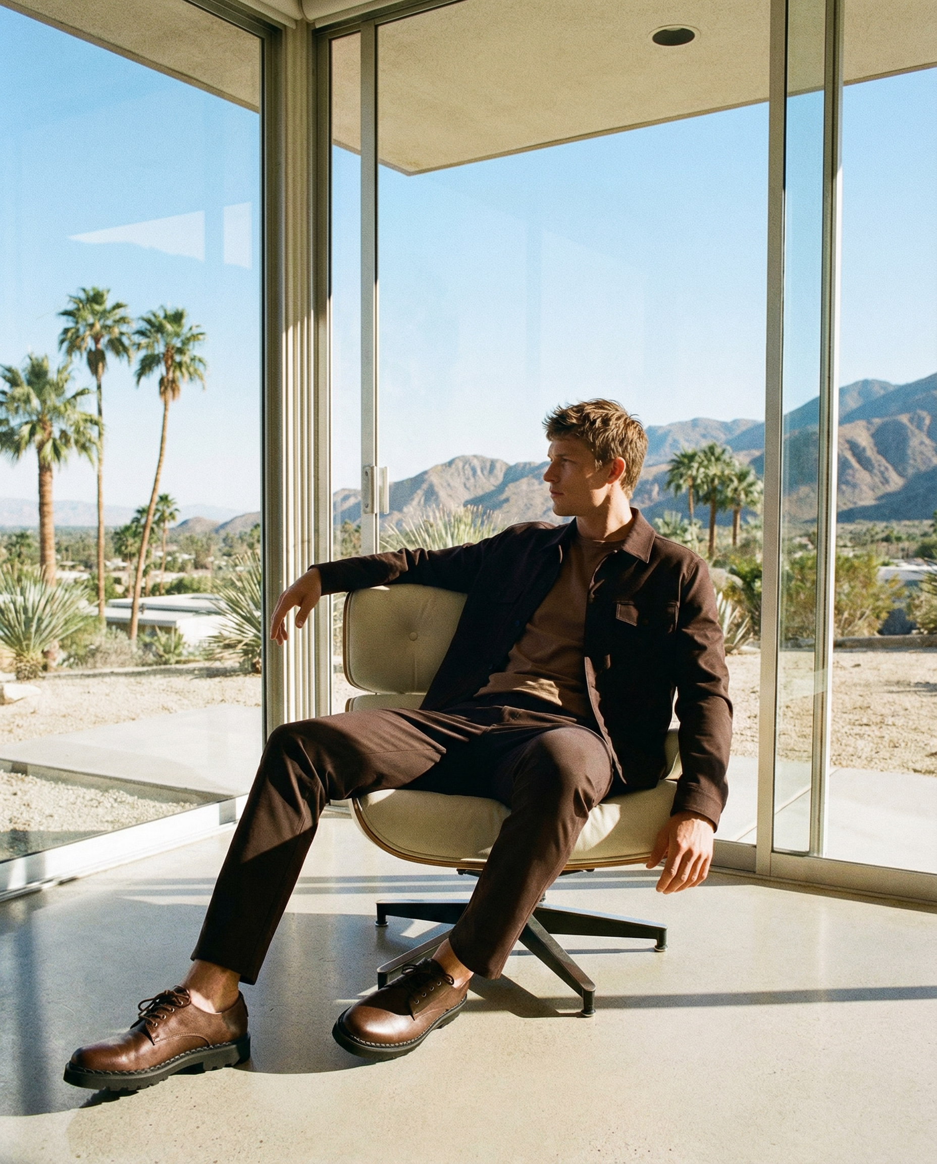 A young man sitting on a modern chair, looking out of a large window at a desert landscape with palm trees, mountains, and clear blue sky. The interior has minimalist decor.