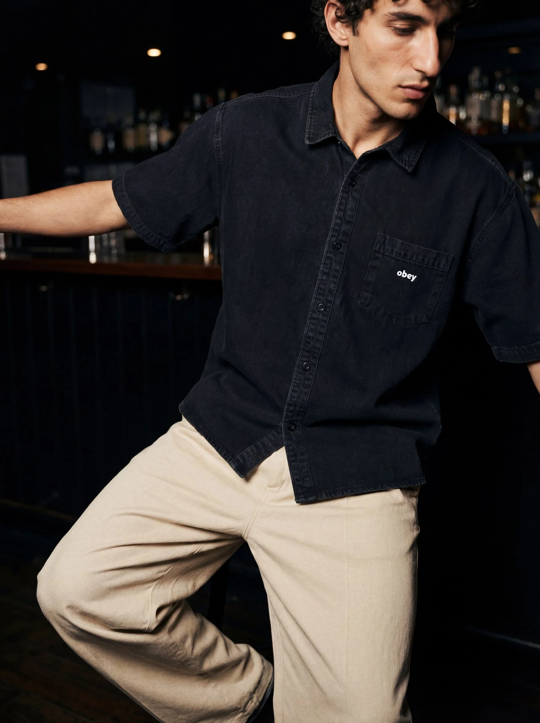 A person wearing a black short-sleeved shirt with 'obey' written on the pocket and beige pants in a dimly lit bar or lounge.