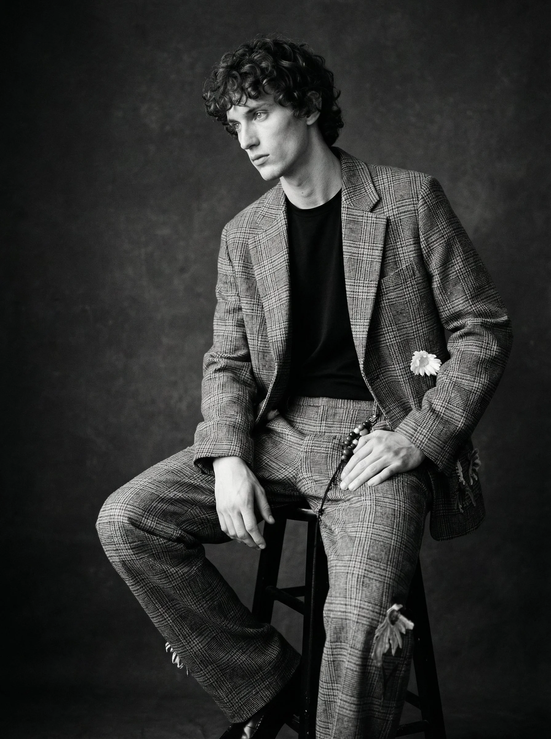Black and white portrait of a young man with curly hair, wearing a checkered blazer and matching pants, sitting on a stool against a dark background, holding a flower and a string of beads.